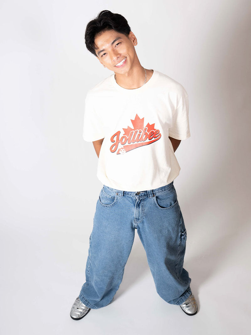 A man wearing a white crewneck t-shirt with a red 'Jolliwise' maple leaf graphic, paired with baggy blue jeans and silver sneakers, posing against a light gray studio background.