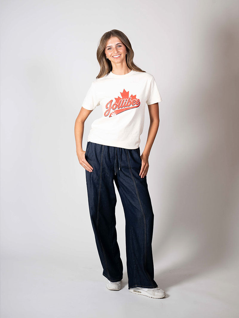 A woman modeling a white crewneck t-shirt with a red 'Jolliets' maple leaf graphic, paired with dark blue wide-leg jeans and white sneakers, against a light gray studio background.