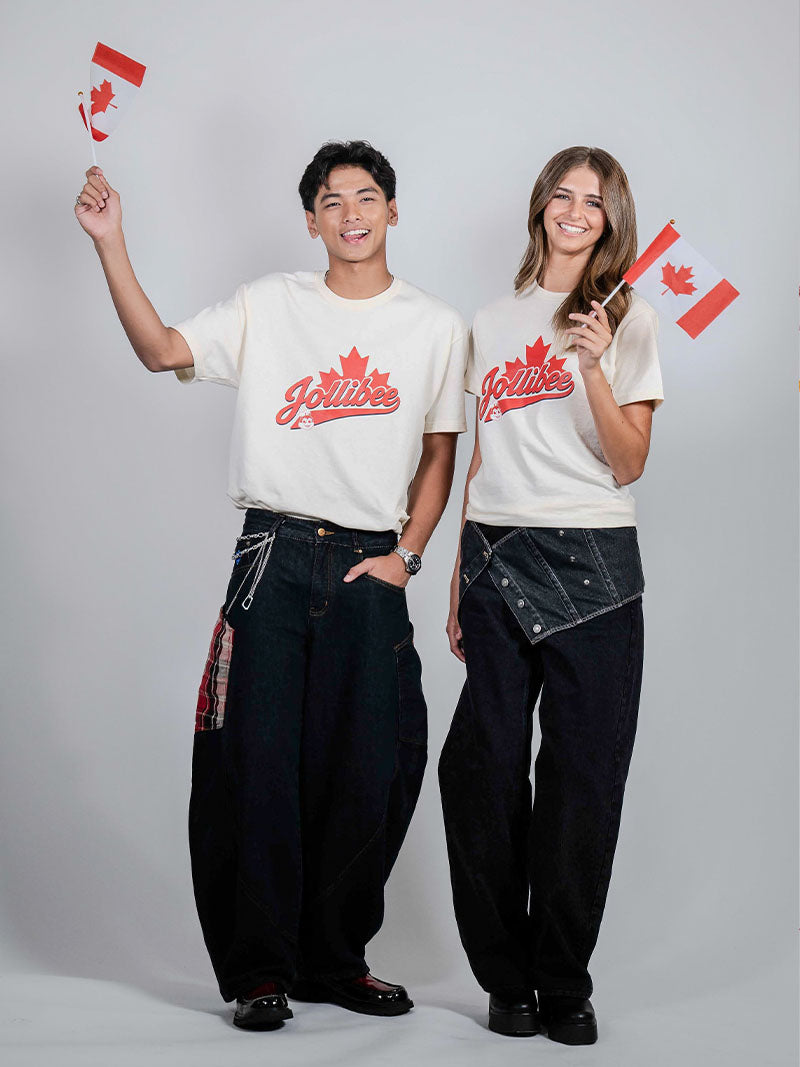 A man and a woman standing side-by-side in a studio, wearing matching white graphic t-shirts with a red 'Vancouver' script and maple leaf, paired with dark baggy jeans. They are holding small Canadian flags against a light gray background.