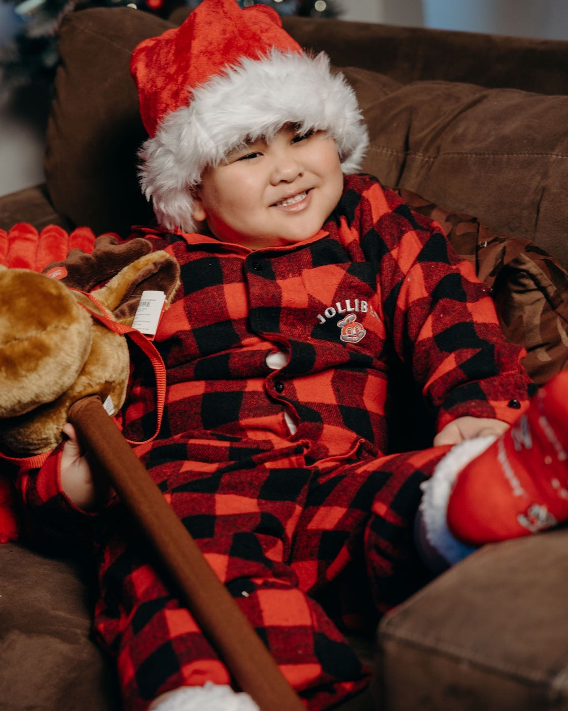 Child in red and black checkered pajamas with Santa hat and mittens, holding a toy, sitting on a brown couch.
