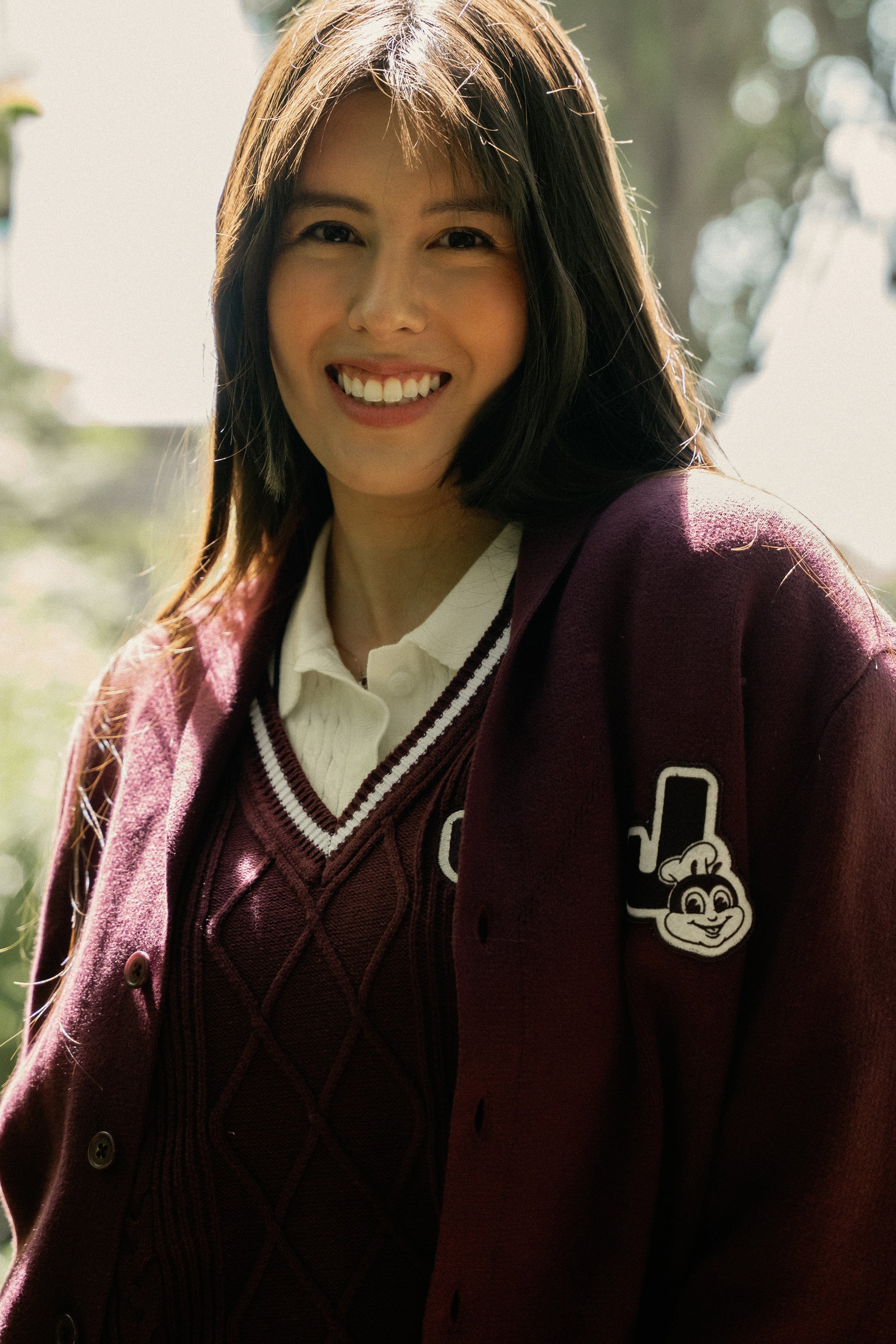 Person wearing a maroon sweater vest and cardigan with a logo, smiling outdoors.