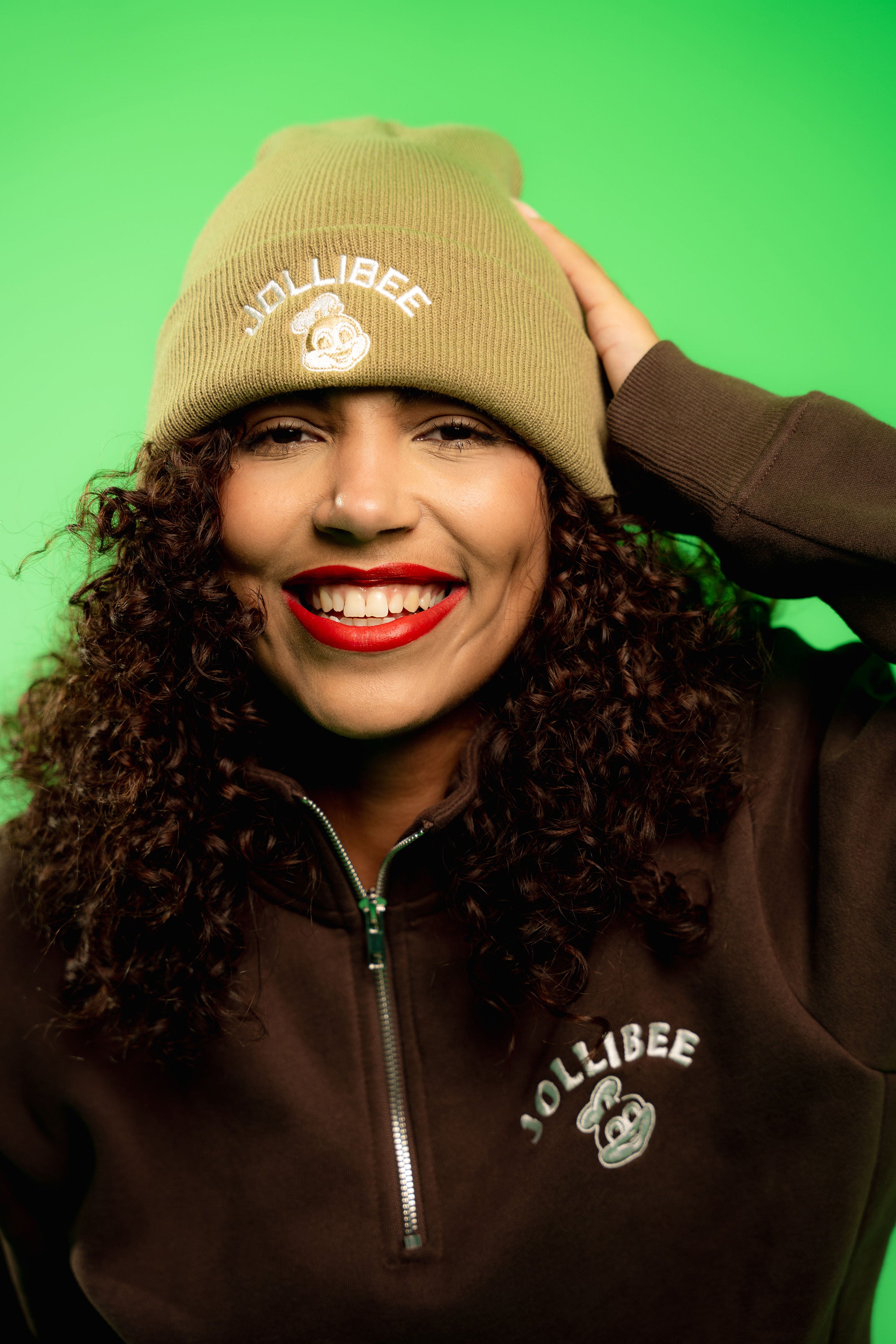 Person wearing a taupe beanie and jacket with 'Jollibee' logo on a green background