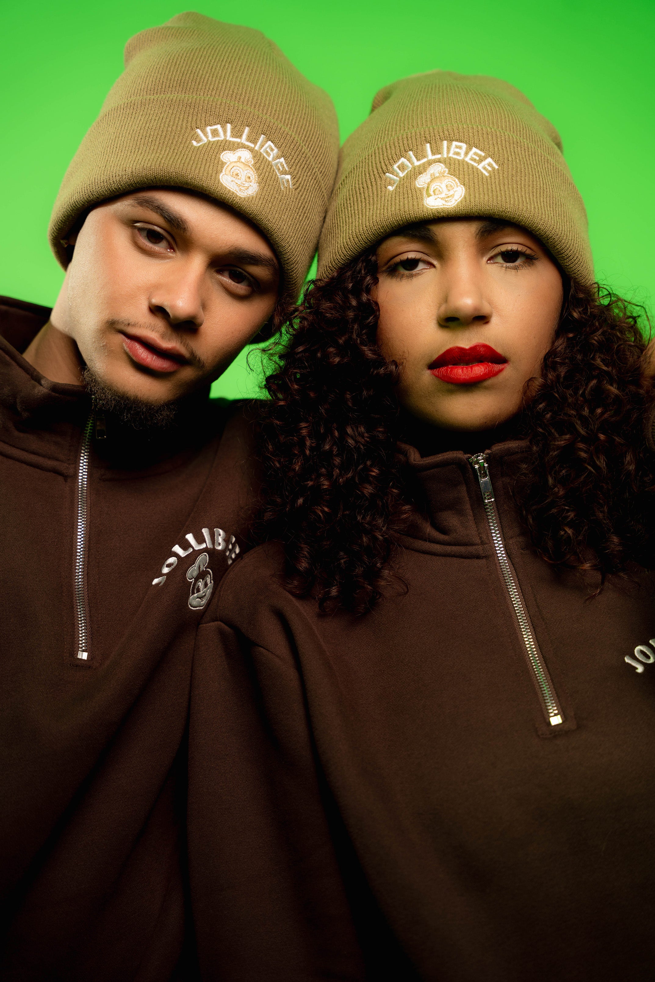 Two people wearing brown jackets and beanie hats with 'Jollibee' branding against a green background.