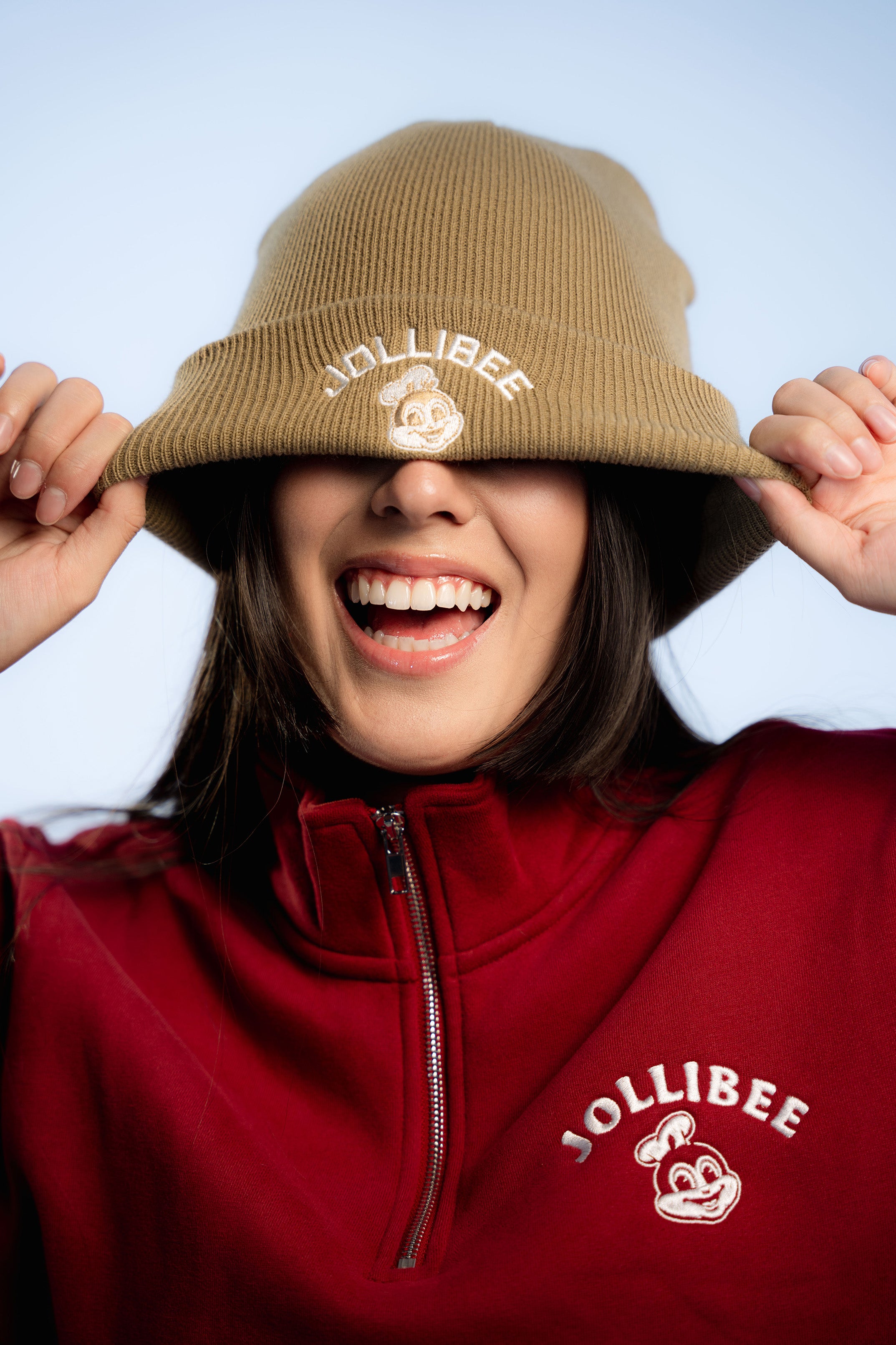 Person wearing a taupe beanie and red zip-up jacket with Jollibee logo on a light blue background