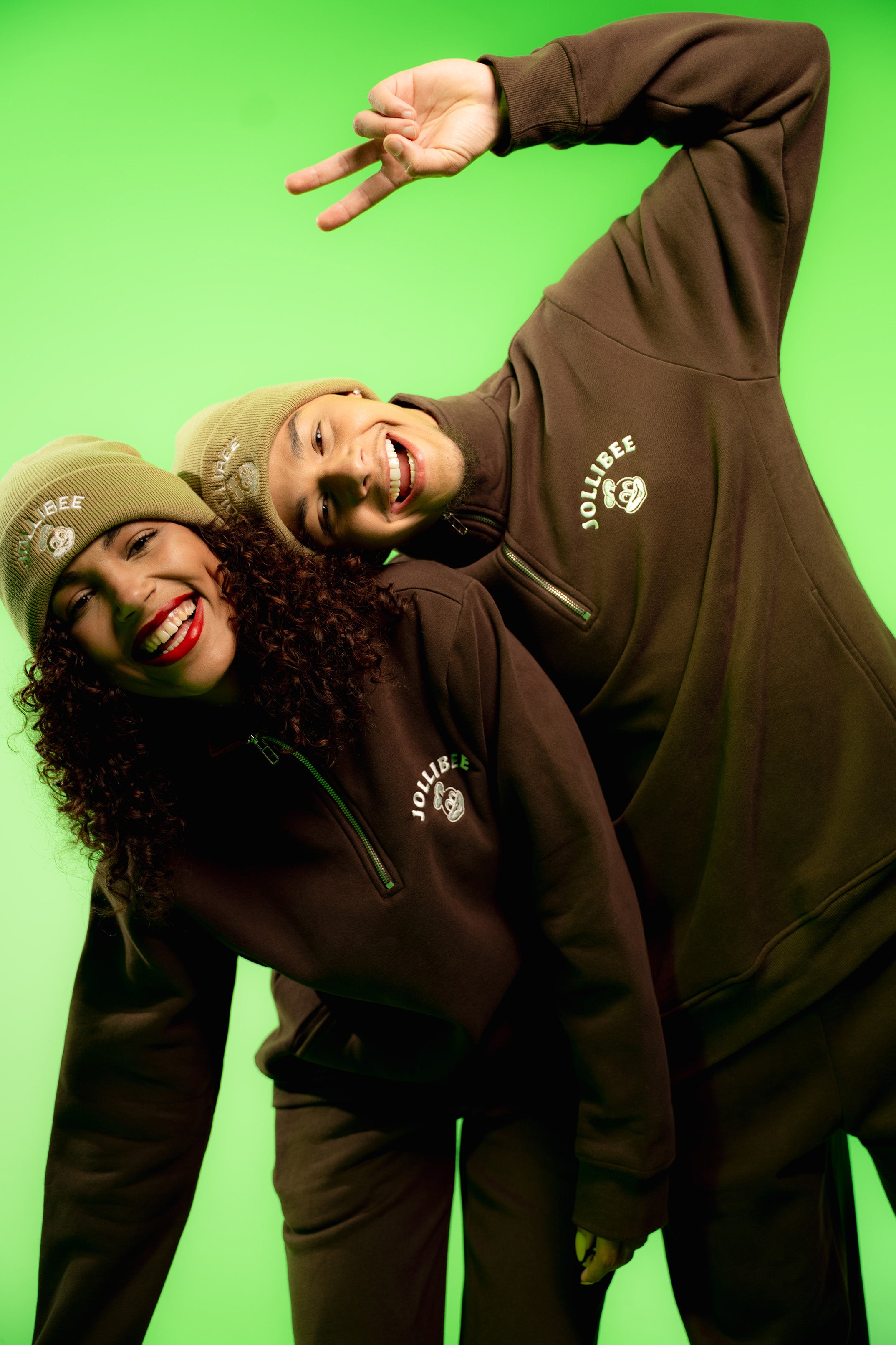 Two people wearing taupe beanies and brown sweatshirts with a logo against a green background