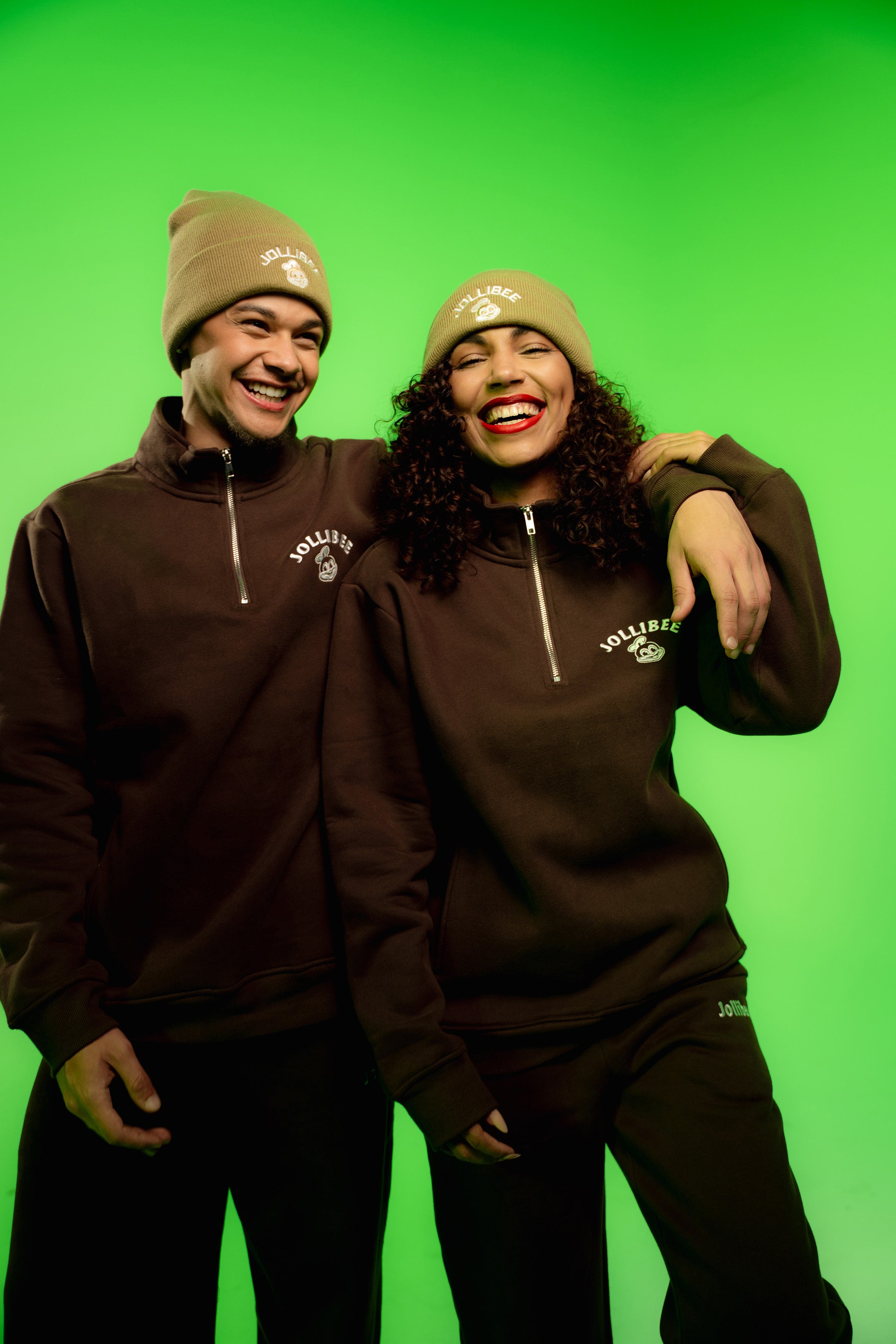 Two people wearing matching brown sweatshirts and beanies against a green background