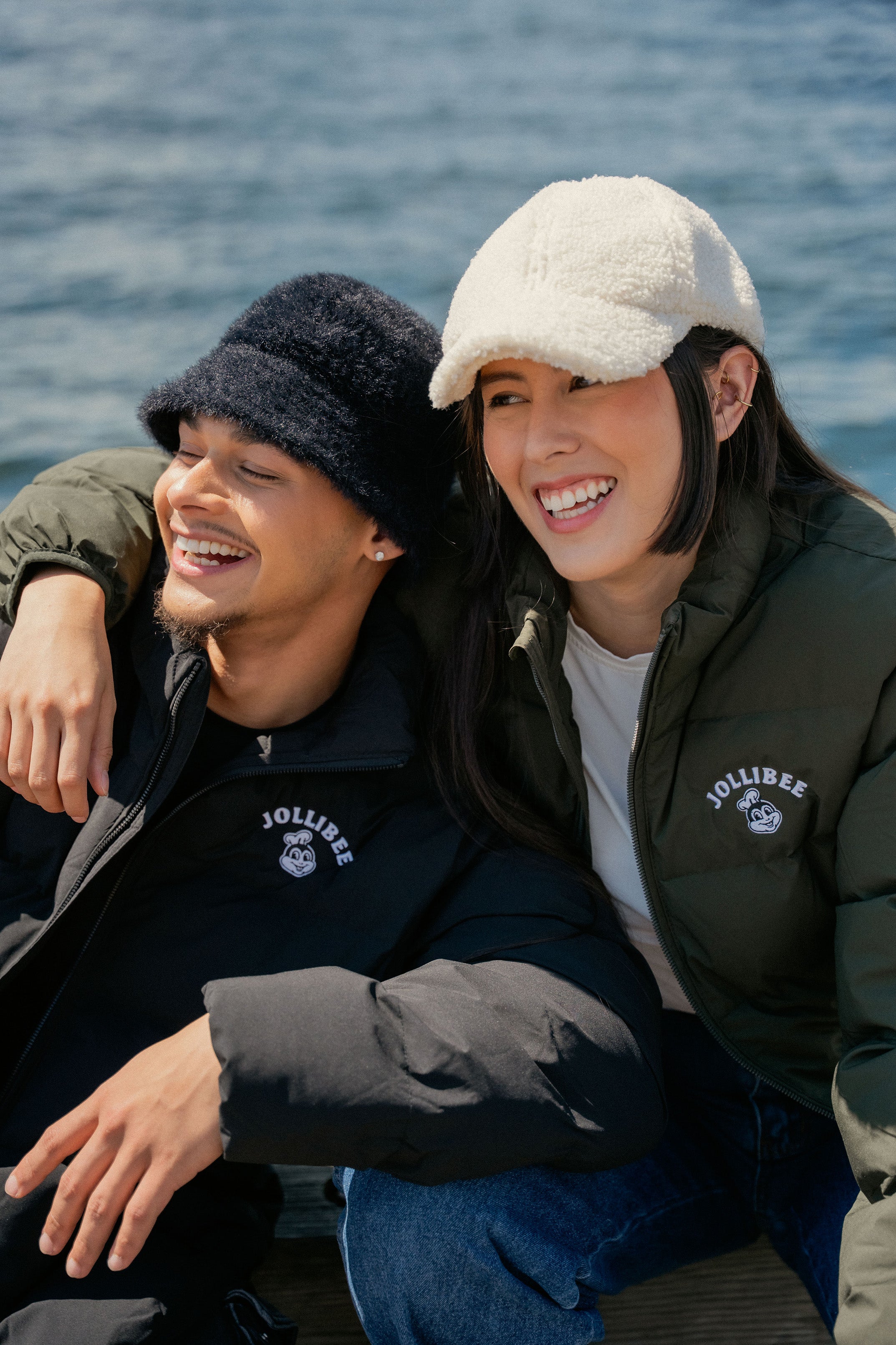 Two people sitting together outdoors with a body of water in the background, wearing Jollibee puffer jackets