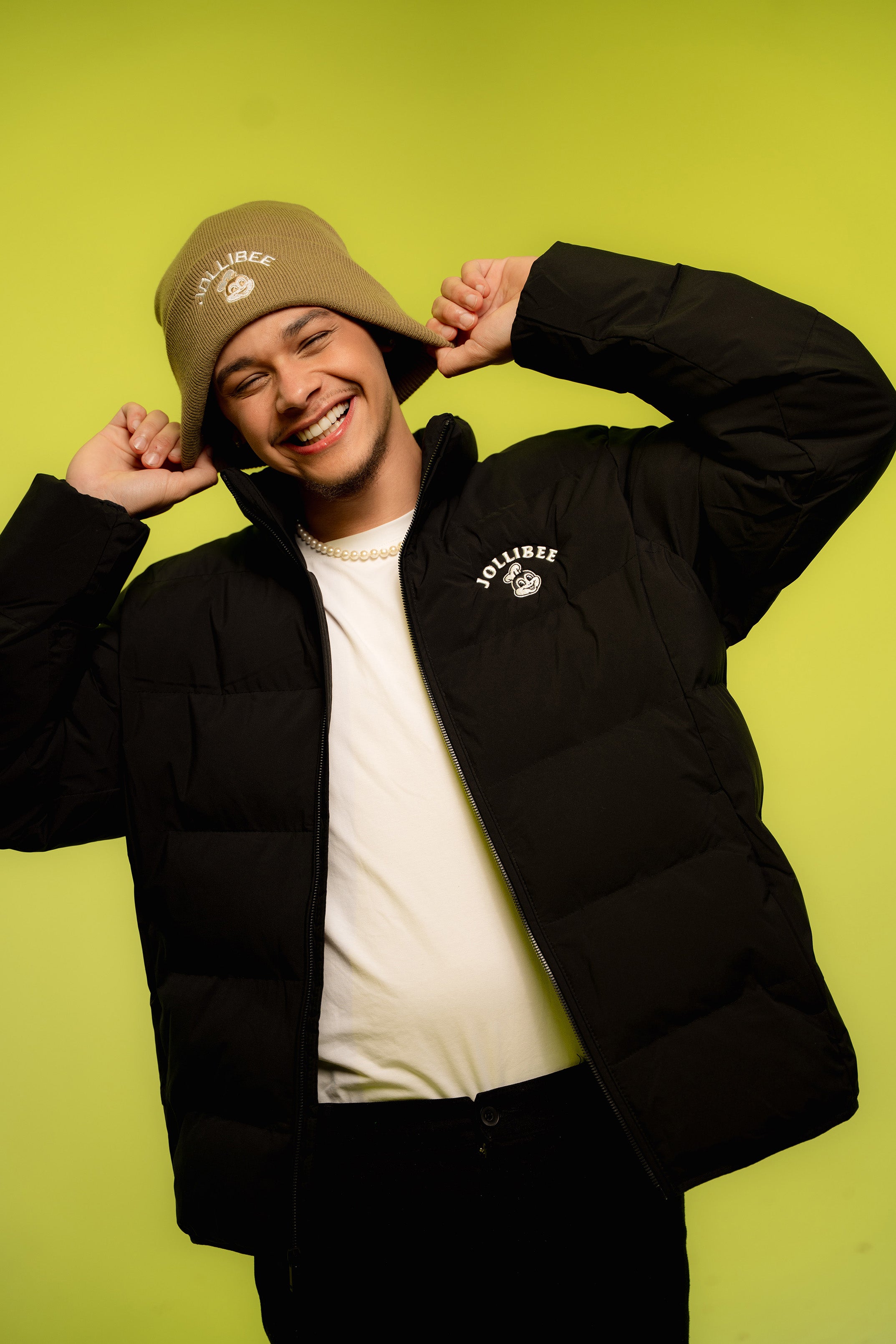 Person wearing a black puffer jacket and taupe beanie against a green background