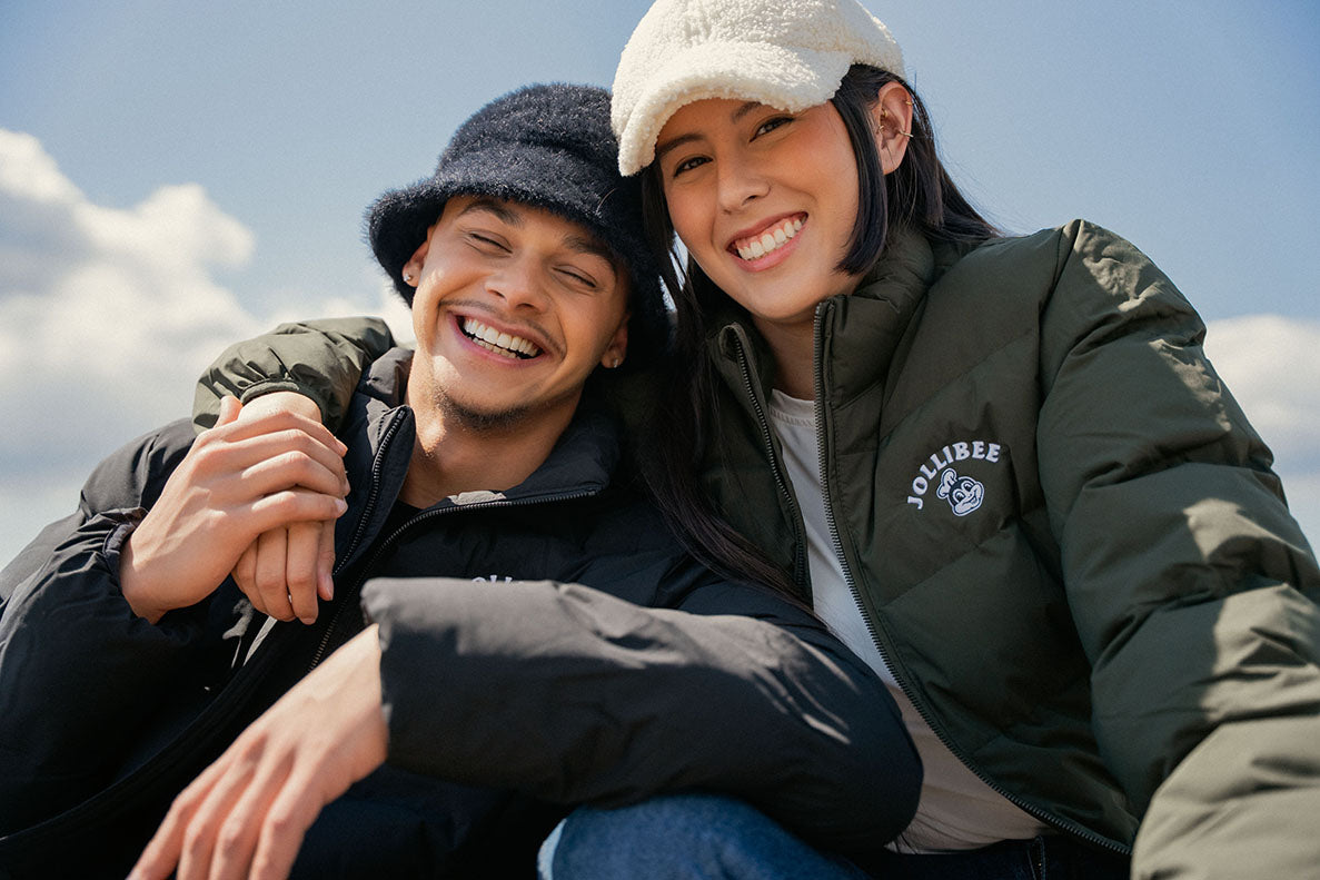 Two people wearing Jollibee jackets and beanies, smiling outdoors with a clear sky.