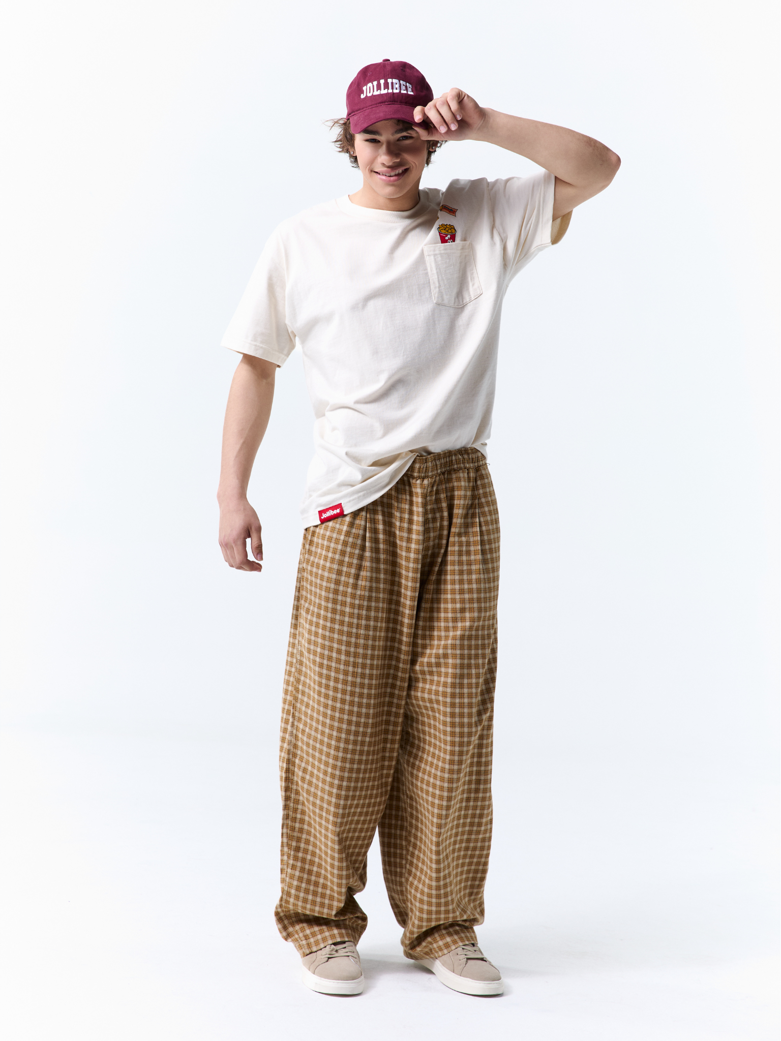 A model wearing a white crewneck t-shirt with a small red chest graphic, brown and beige checkered wide-leg pants, a burgundy baseball cap, and white sneakers, posing against a white studio background.