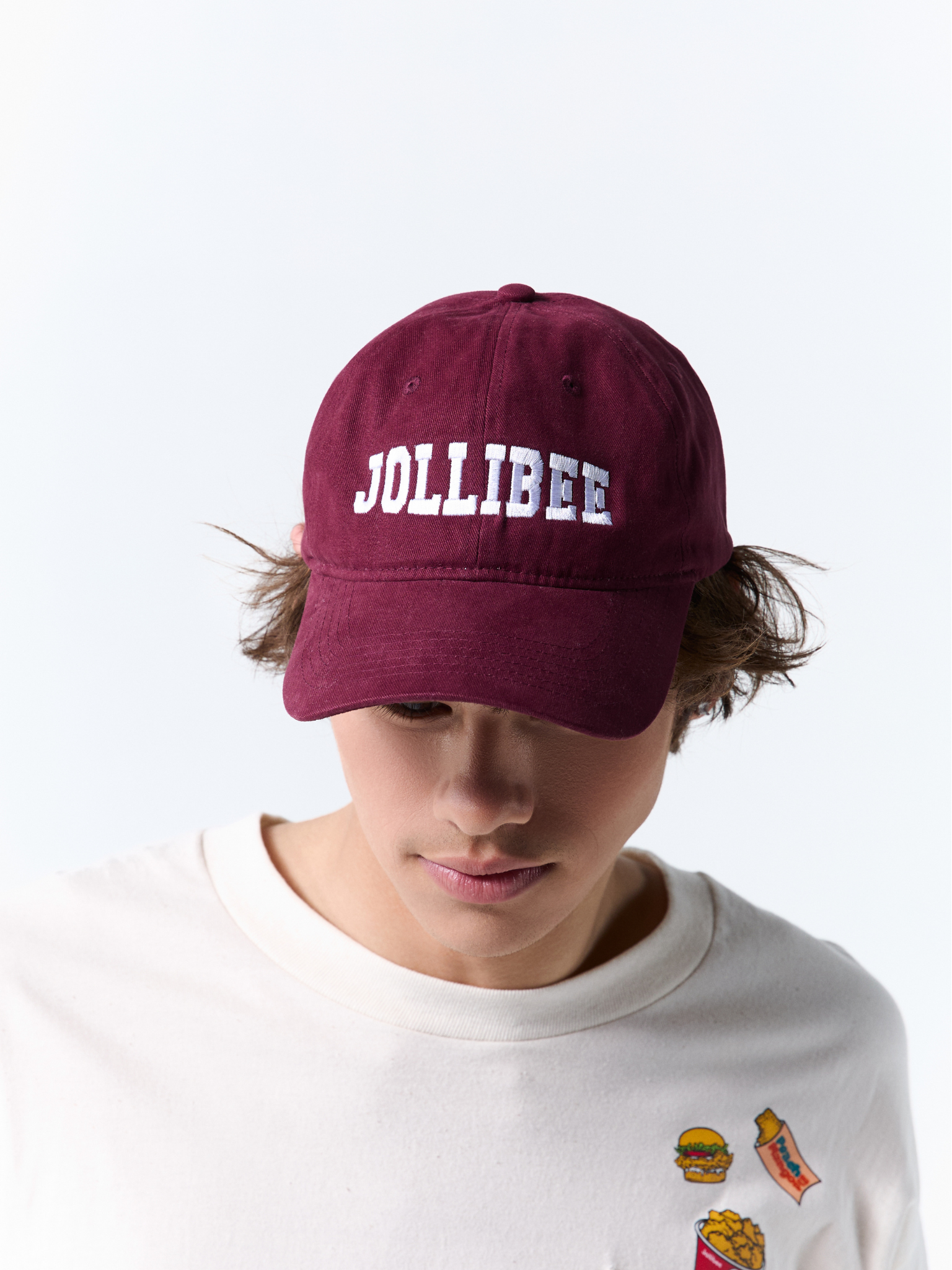 A person wearing a maroon baseball cap with 'JOLLIBEE' embroidered in white across the front, paired with a white graphic t-shirt against a plain white background.