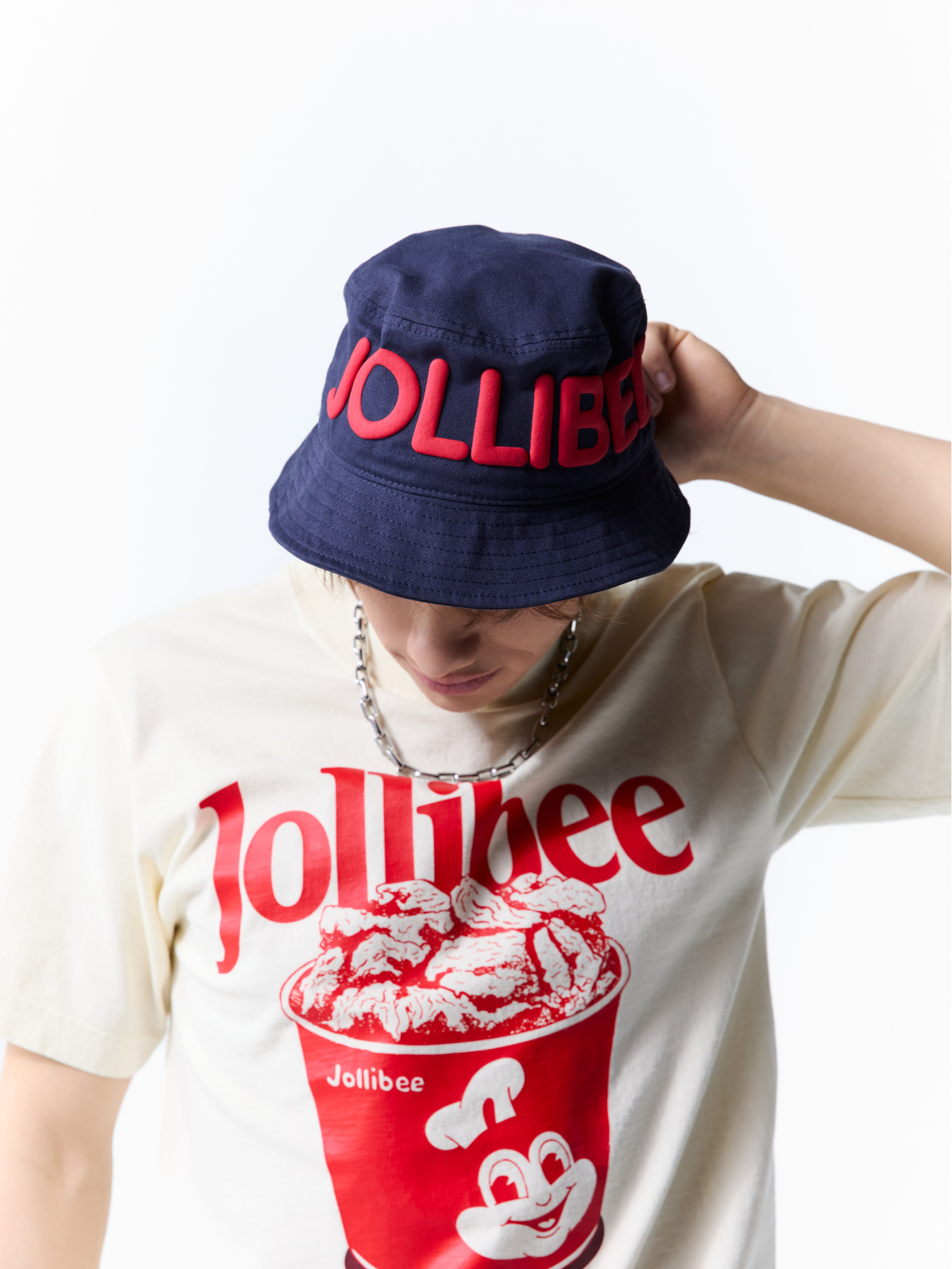 A person wearing a navy blue Jollibee bucket hat with red embroidered lettering and a cream-colored Jollibee graphic t-shirt, shown against a plain white background.