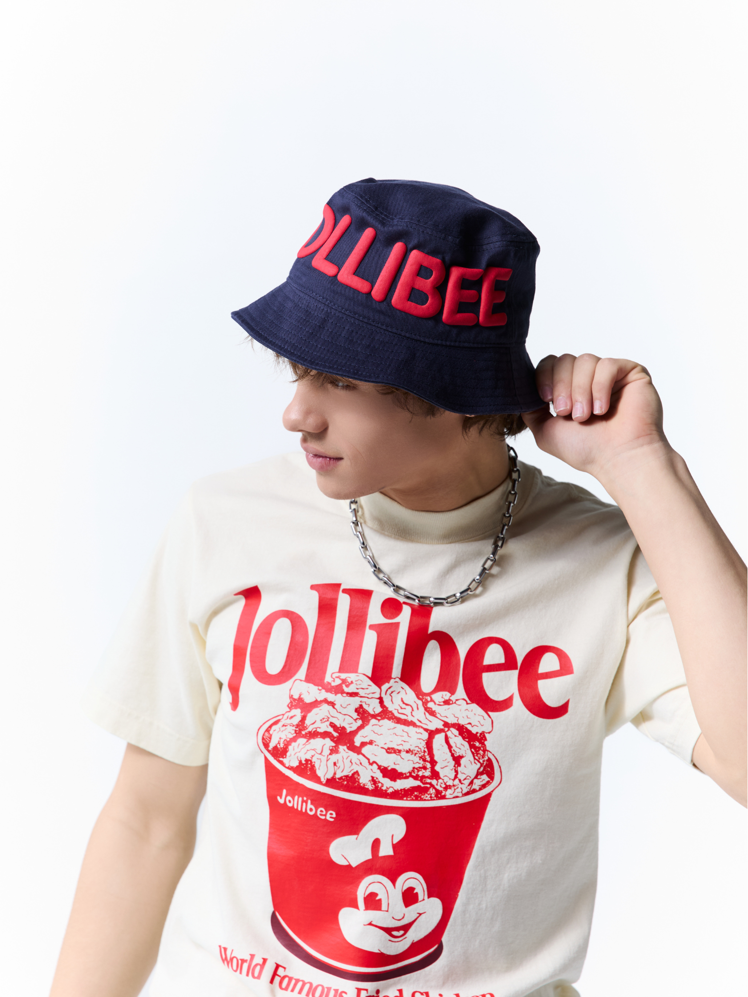 A person wearing a navy blue Jollibee bucket hat with red lettering and a cream-colored Jollibee graphic t-shirt, set against a plain white background.