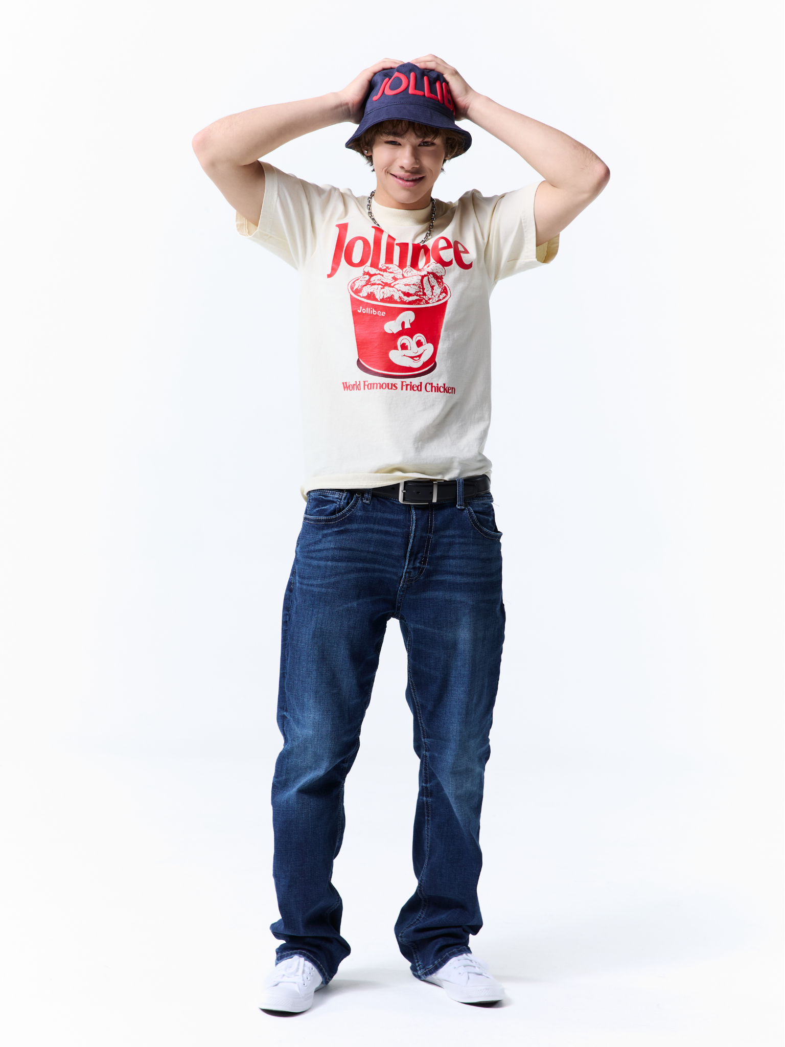 A person modeling a cream graphic t-shirt with a red Jollibee bucket print, paired with dark blue jeans, white sneakers, and a navy blue bucket hat with red lettering, against a plain white background.