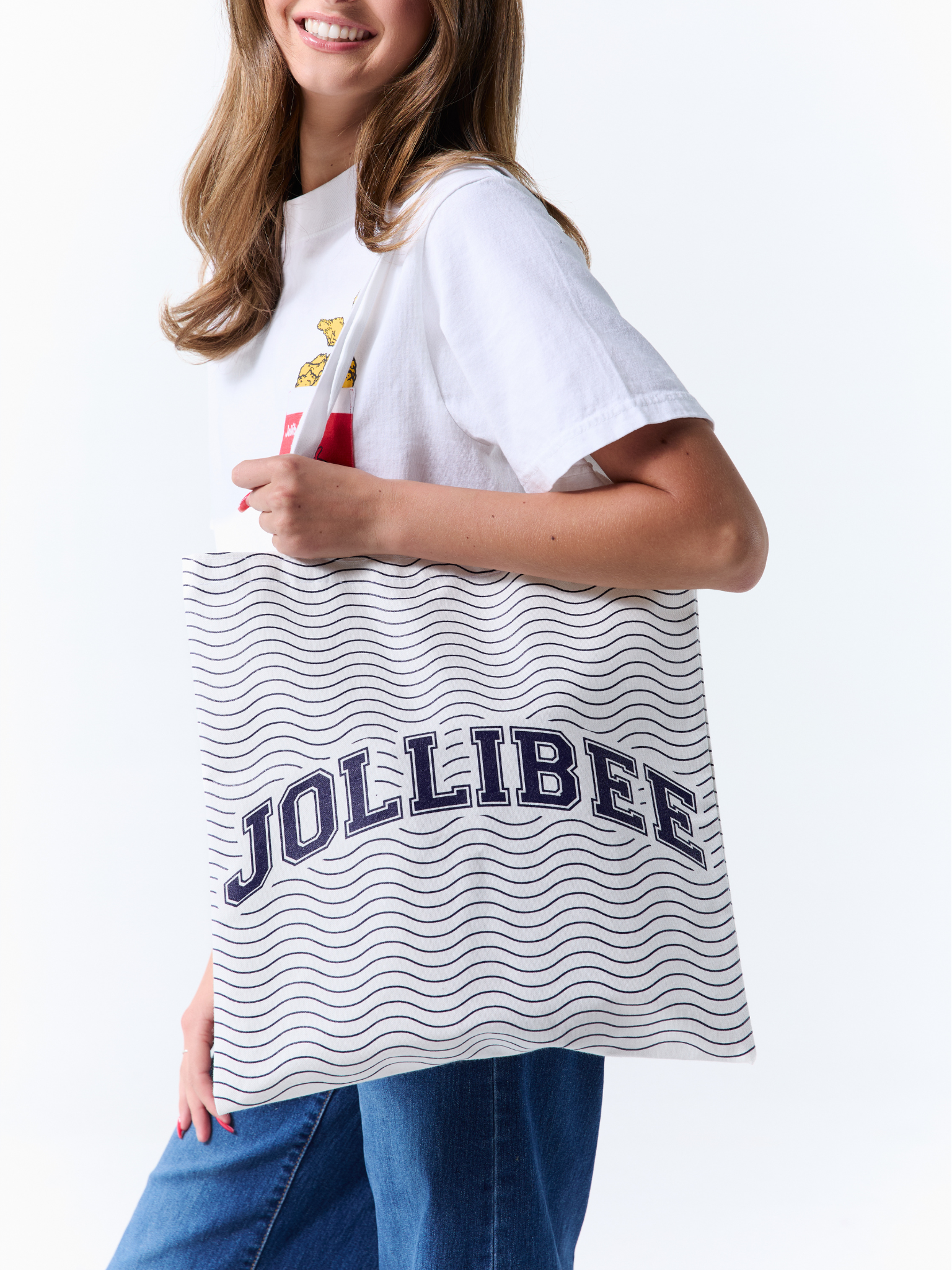 A woman wearing a white graphic t-shirt and blue jeans carries a white canvas tote bag with navy blue wavy stripes and the word 'JOLLIBEE' printed across the front.