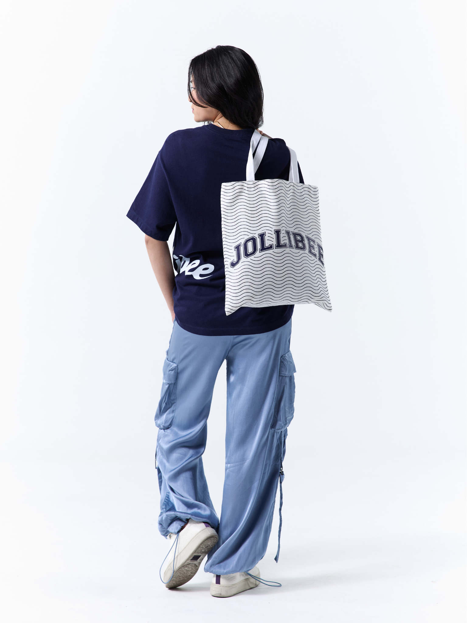 A model is shown from behind wearing a navy blue t-shirt, light blue cargo pants, and white sneakers, while carrying a white tote bag with black wavy stripes and the text 'JOLLIBEE' on the back.