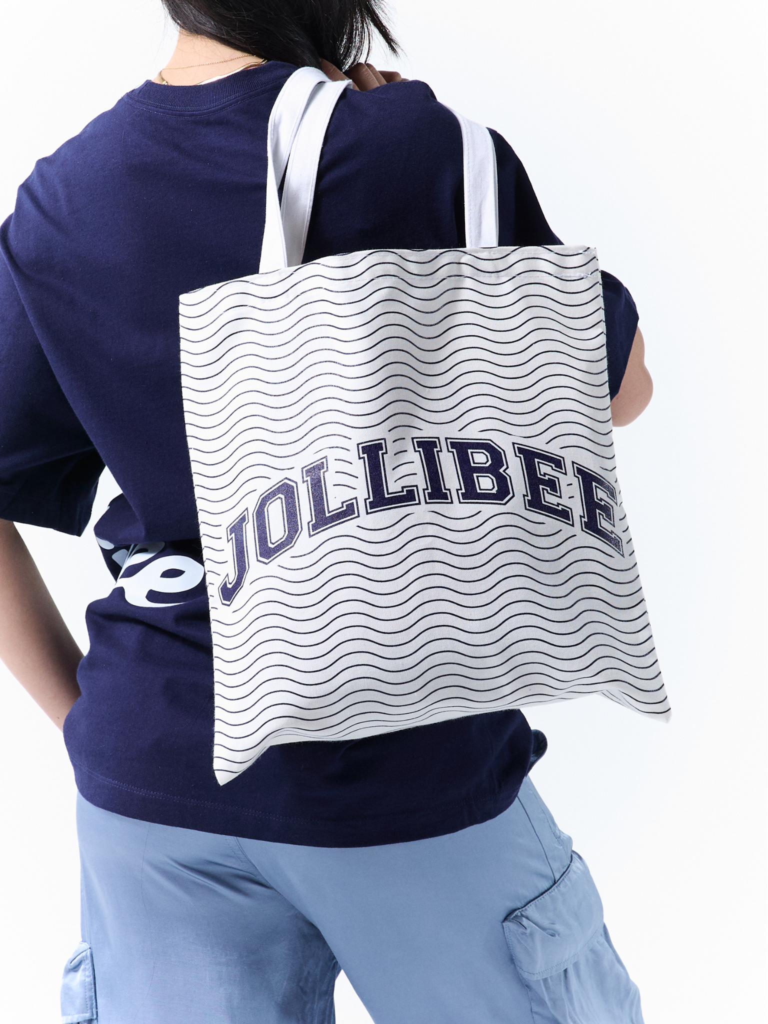 A person shown from the back wearing a navy blue t-shirt and light blue cargo pants, carrying a white tote bag with navy blue wavy stripes and the word 'JOLLIBEE' printed across the front.