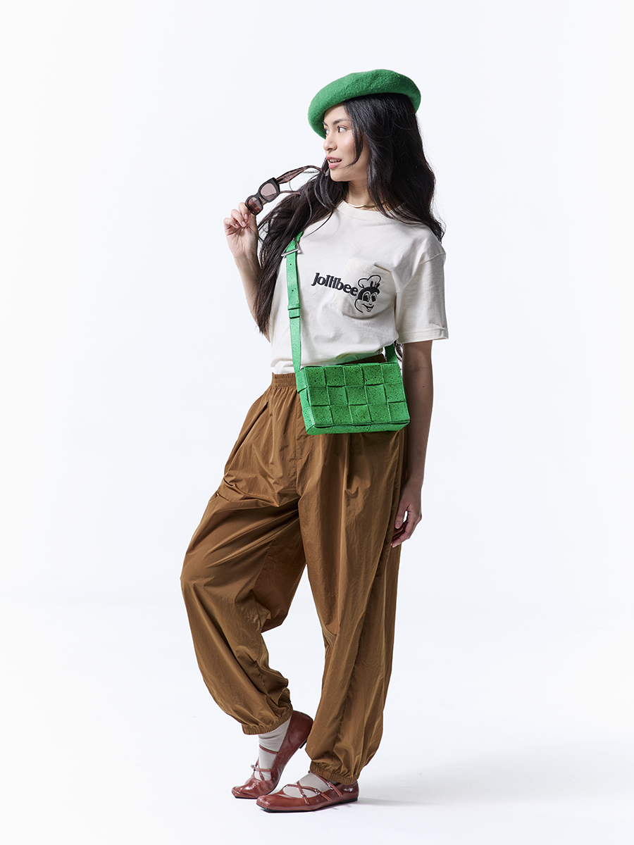 Person wearing a green beret, beige t-shirt with text, brown pants, and a green bag on a white background