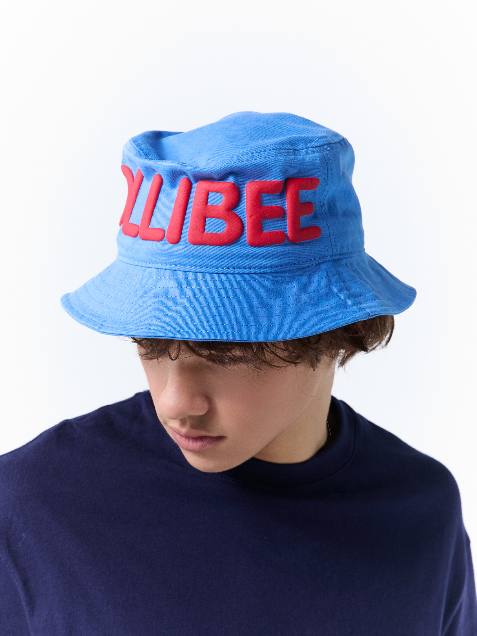 A person wearing a bright blue bucket hat with large red 3D text reading 'JOLLIBEE' on the front, paired with a dark navy shirt against a plain white background.