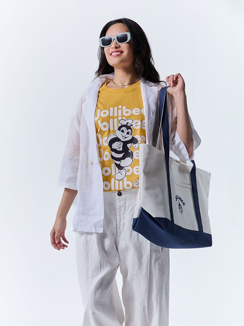 Person wearing a yellow t-shirt with a logo and white pants, holding a tote bag.