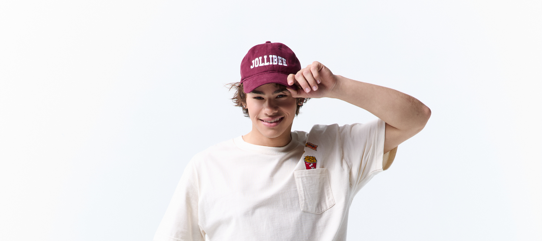 A smiling young man wearing a maroon Jollibee baseball cap and an off-white t-shirt with a chest pocket, posing against a solid white background.