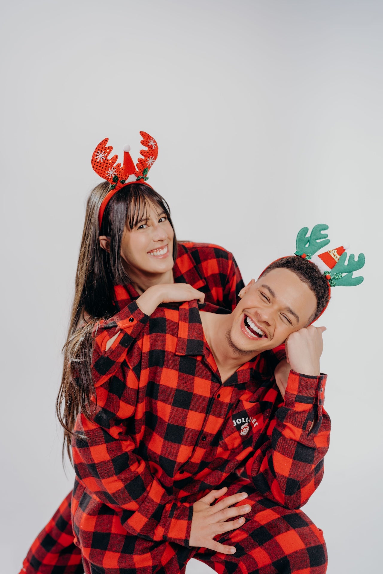 Two people wearing red and black checkered outfits with reindeer antlers on a light gray background.