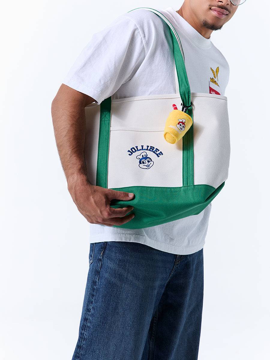 A man wearing a white t-shirt and blue jeans carries a white tote bag with green straps and bottom panel. The bag features a front pocket with 'JOLLIBEE' embroidered text and a small yellow Jollibee pineapple quencher charm.