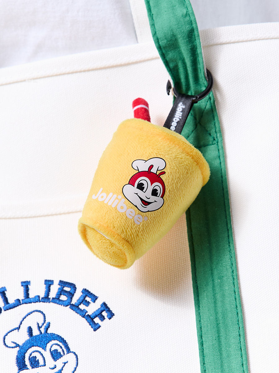 A yellow plush Jollibee pineapple quencher keychain or bag charm clipped to a white tote bag with a green strap. The cup features the Jollibee mascot face and the word 'Jollibee' in white.