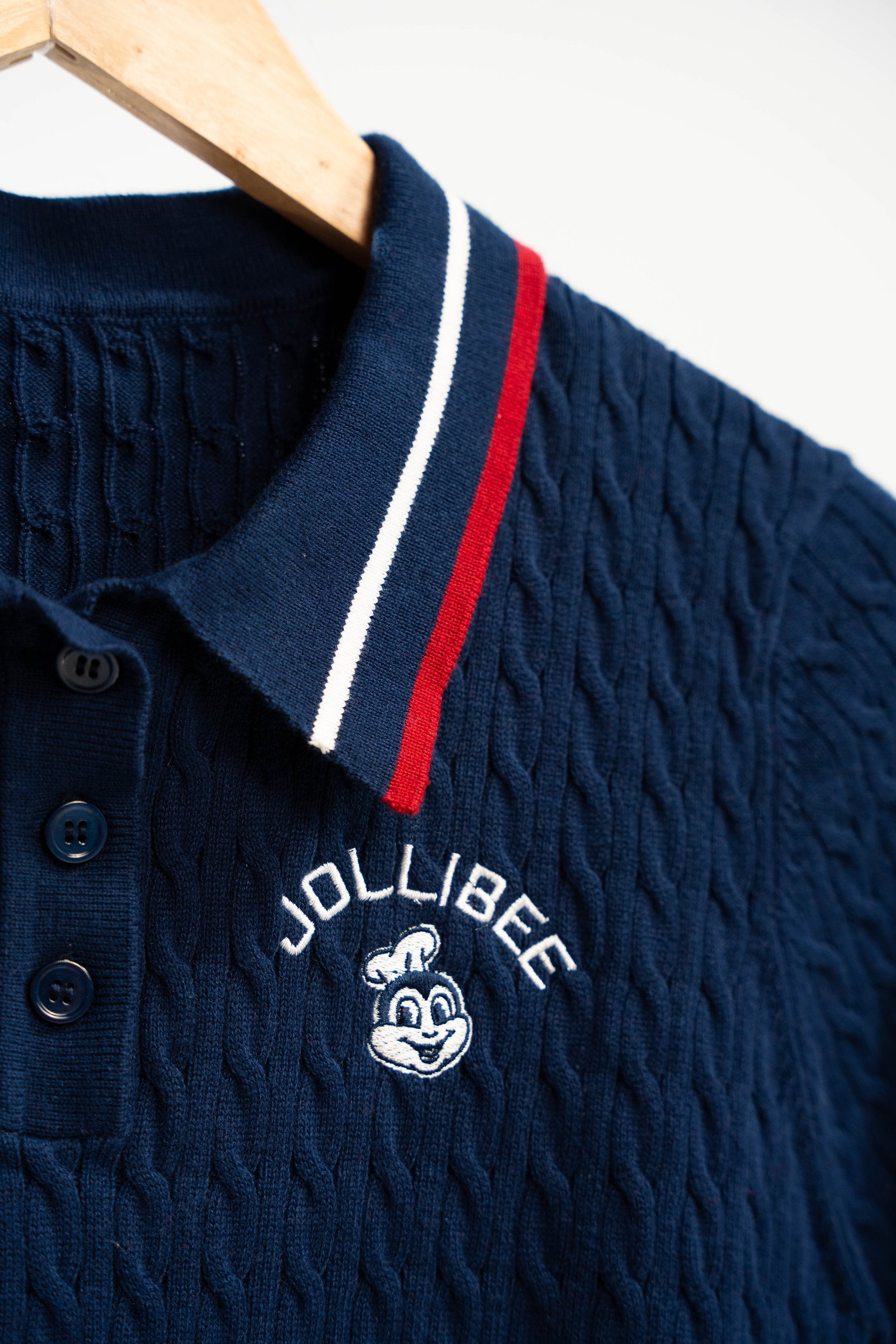 Navy blue textured polo shirt with red and white stripe collar on a hanger, featuring Jollibee logo.