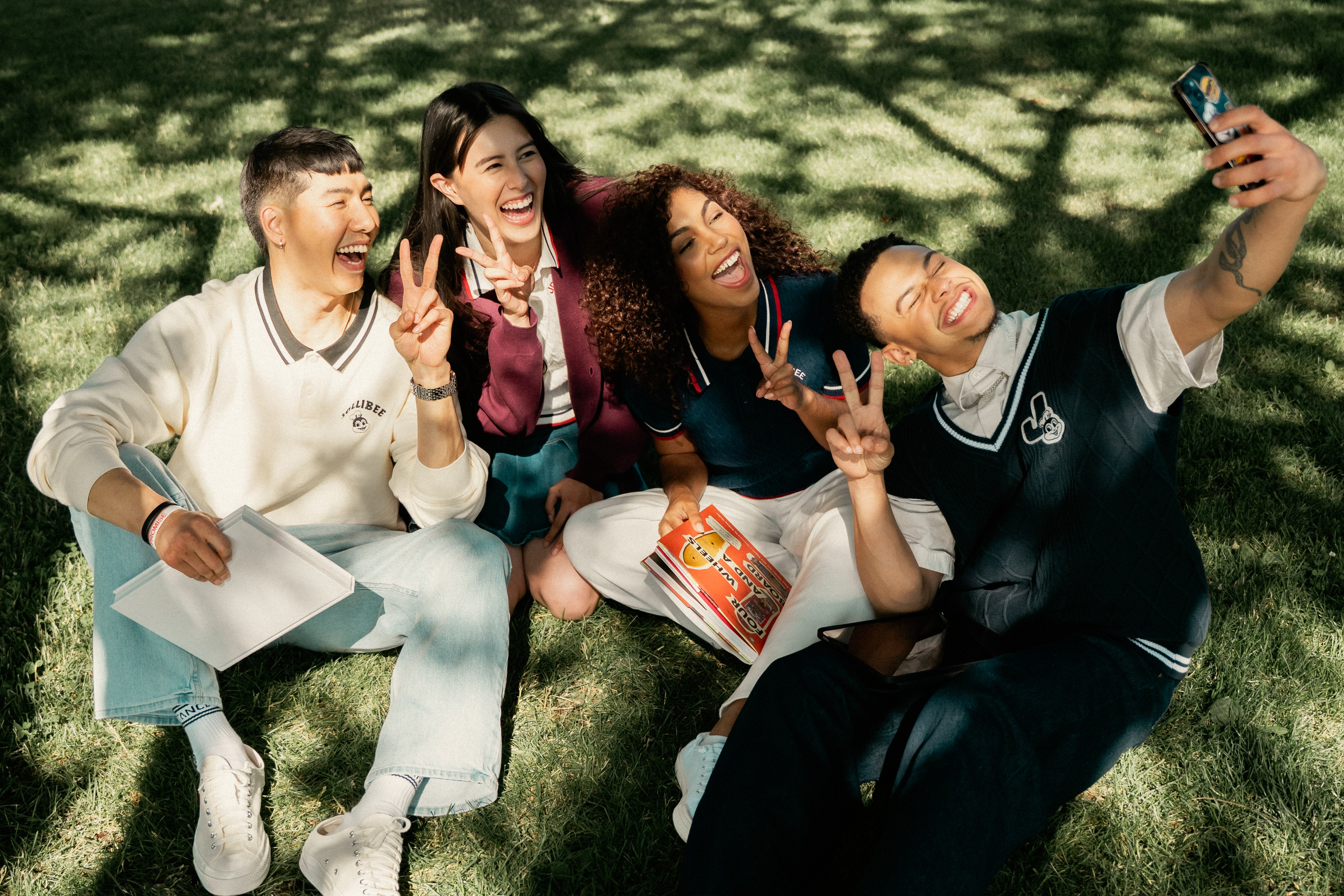 Four friends sitting on grass, taking a selfie and making peace signs.