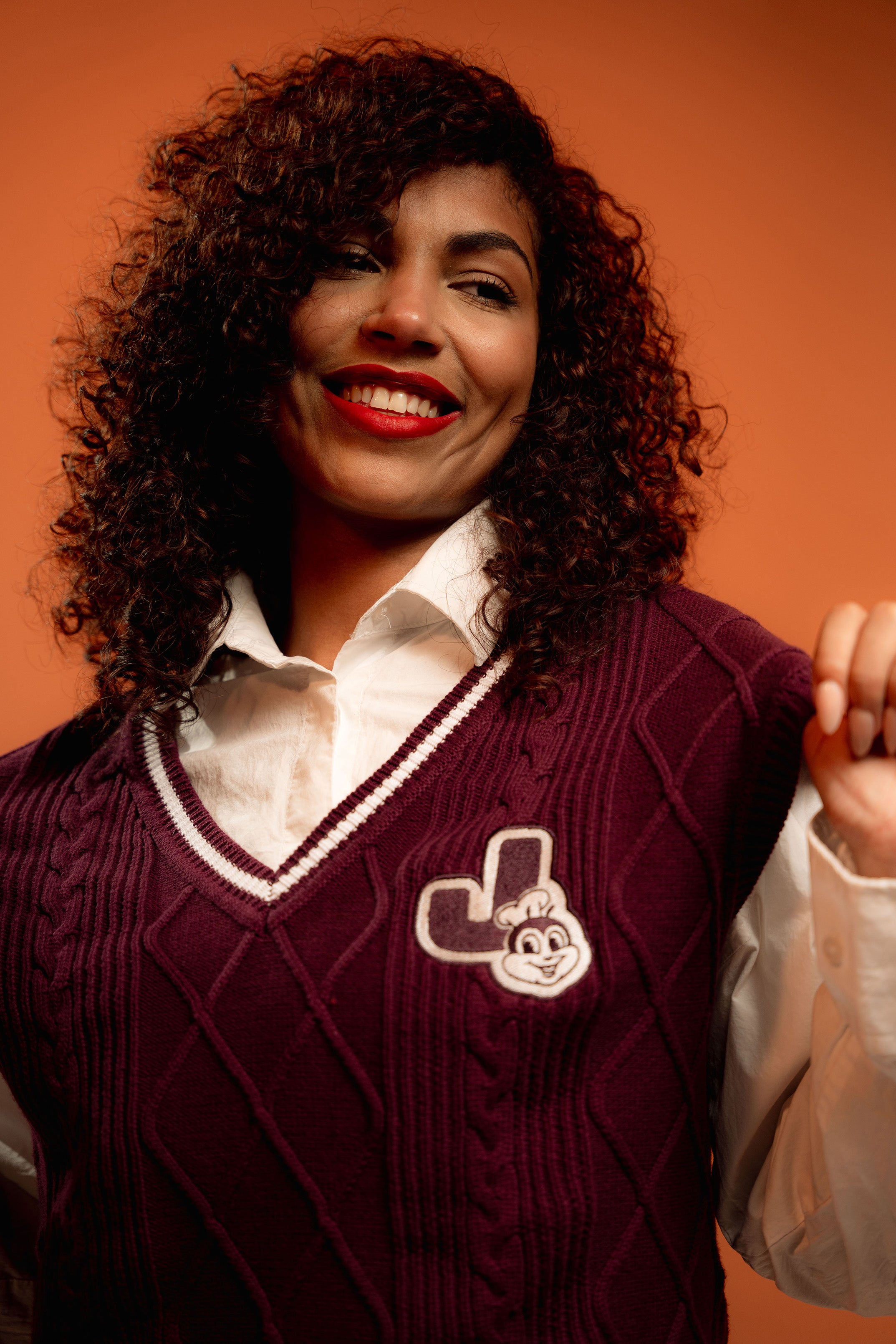 Woman wearing a maroon sweater vest with a logo against an orange background