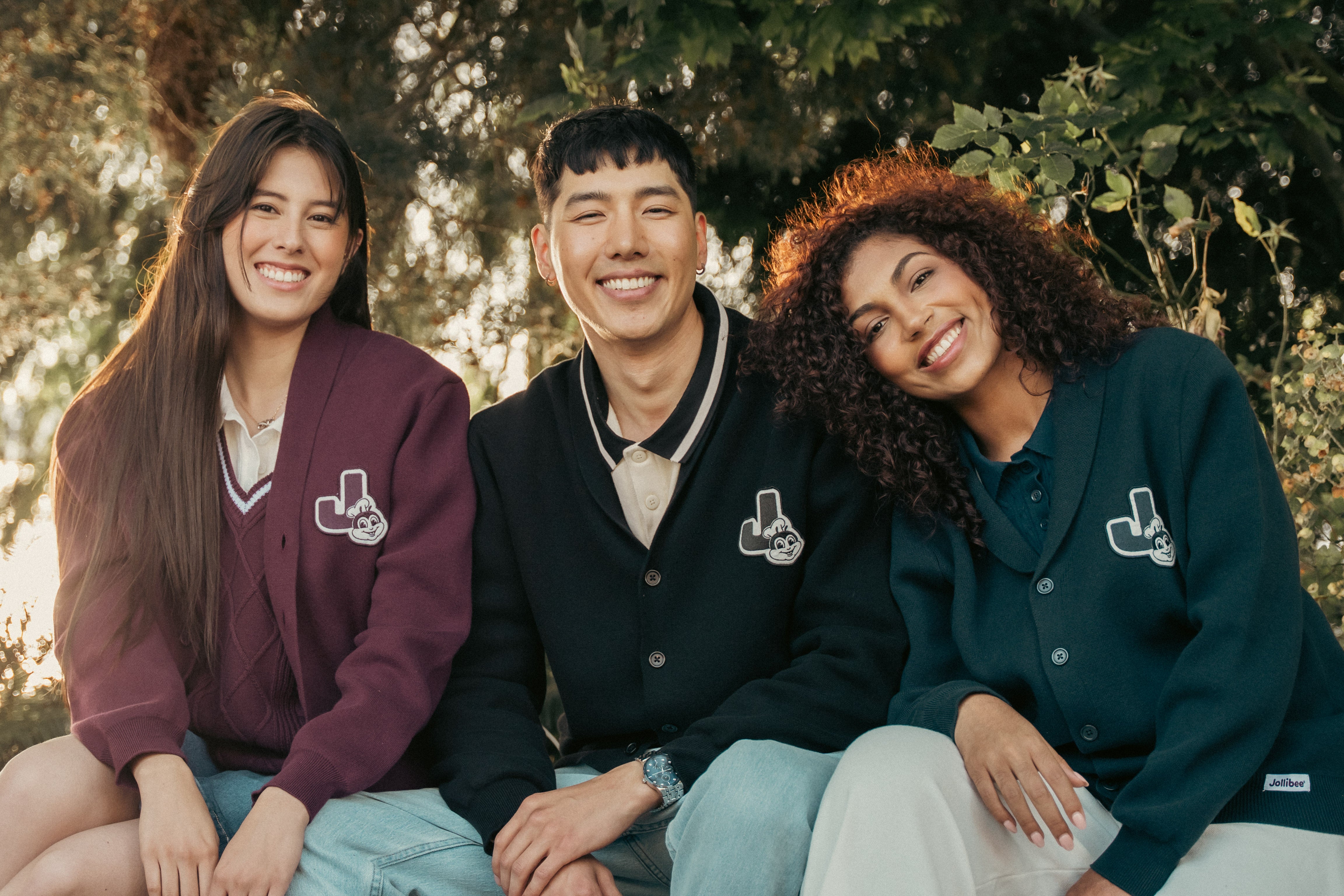 Three people wearing matching Jollibee cardigans sitting outdoors with trees in the background