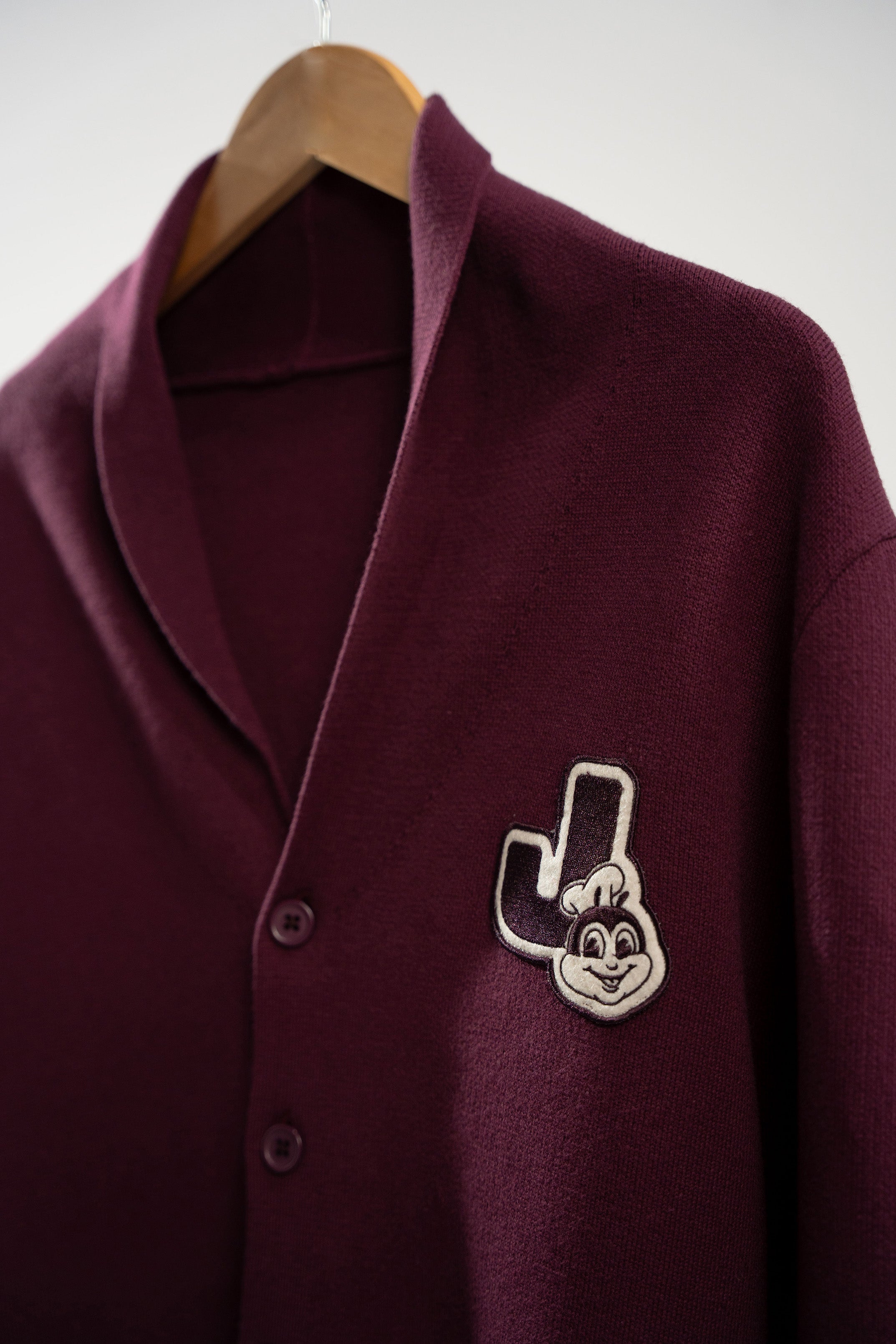 Maroon cardigan with a logo patch on a wooden hanger against a white background