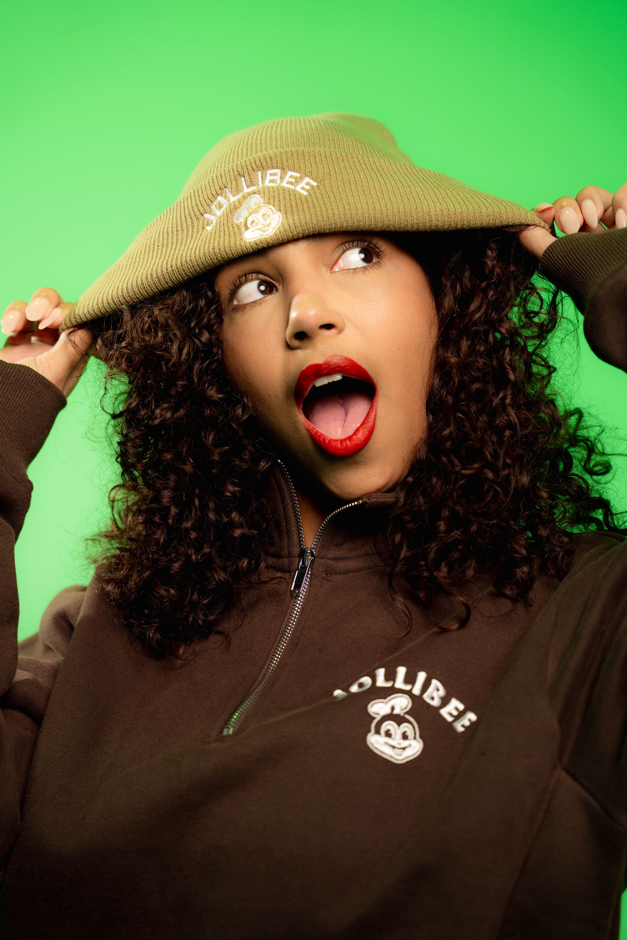 Person wearing a taupe beanie and shirt with 'JOLLIBEE' logo against a green background