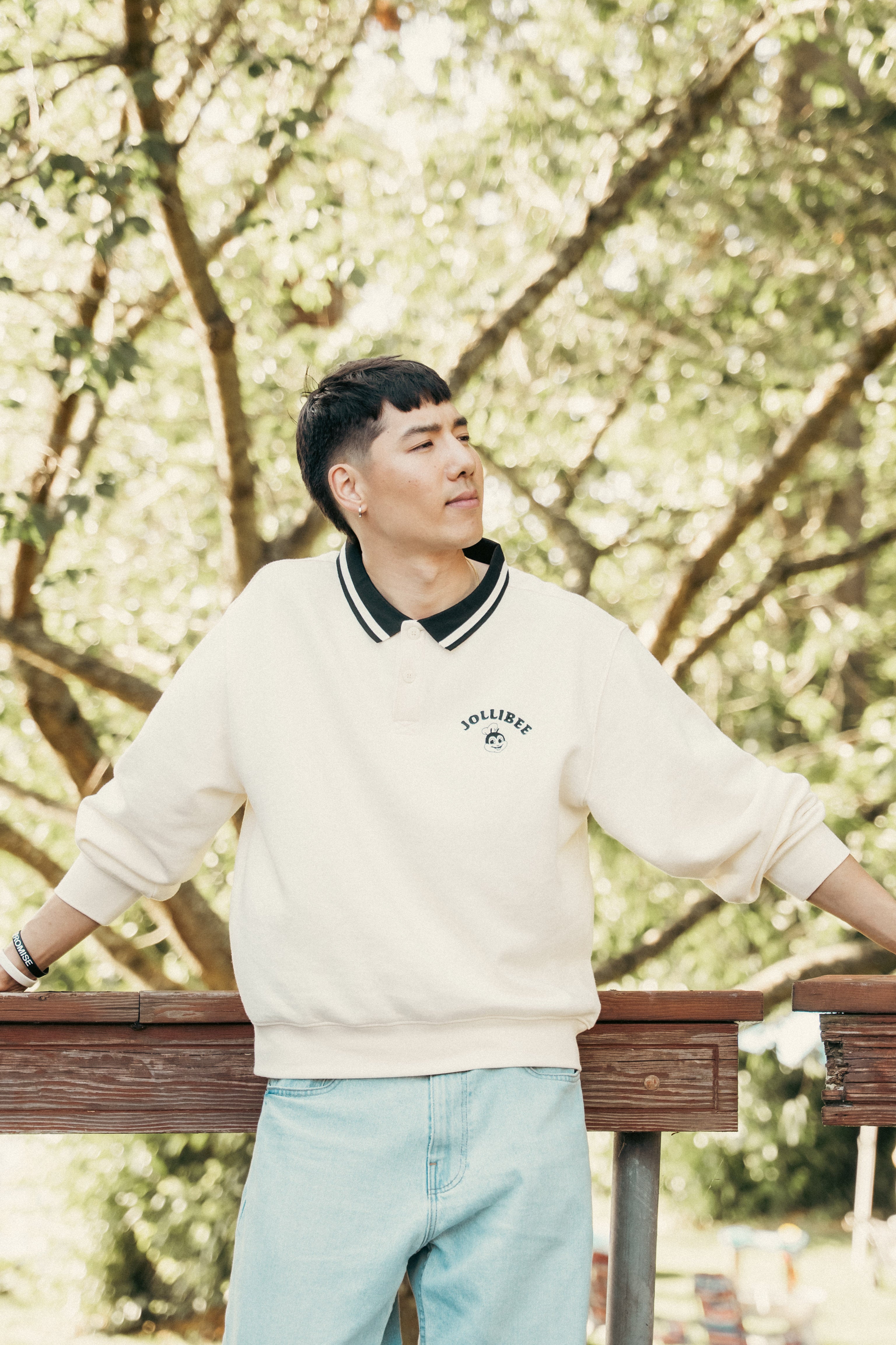 Man wearing a white sweater with a Jollibee logo, standing outdoors with trees in the background