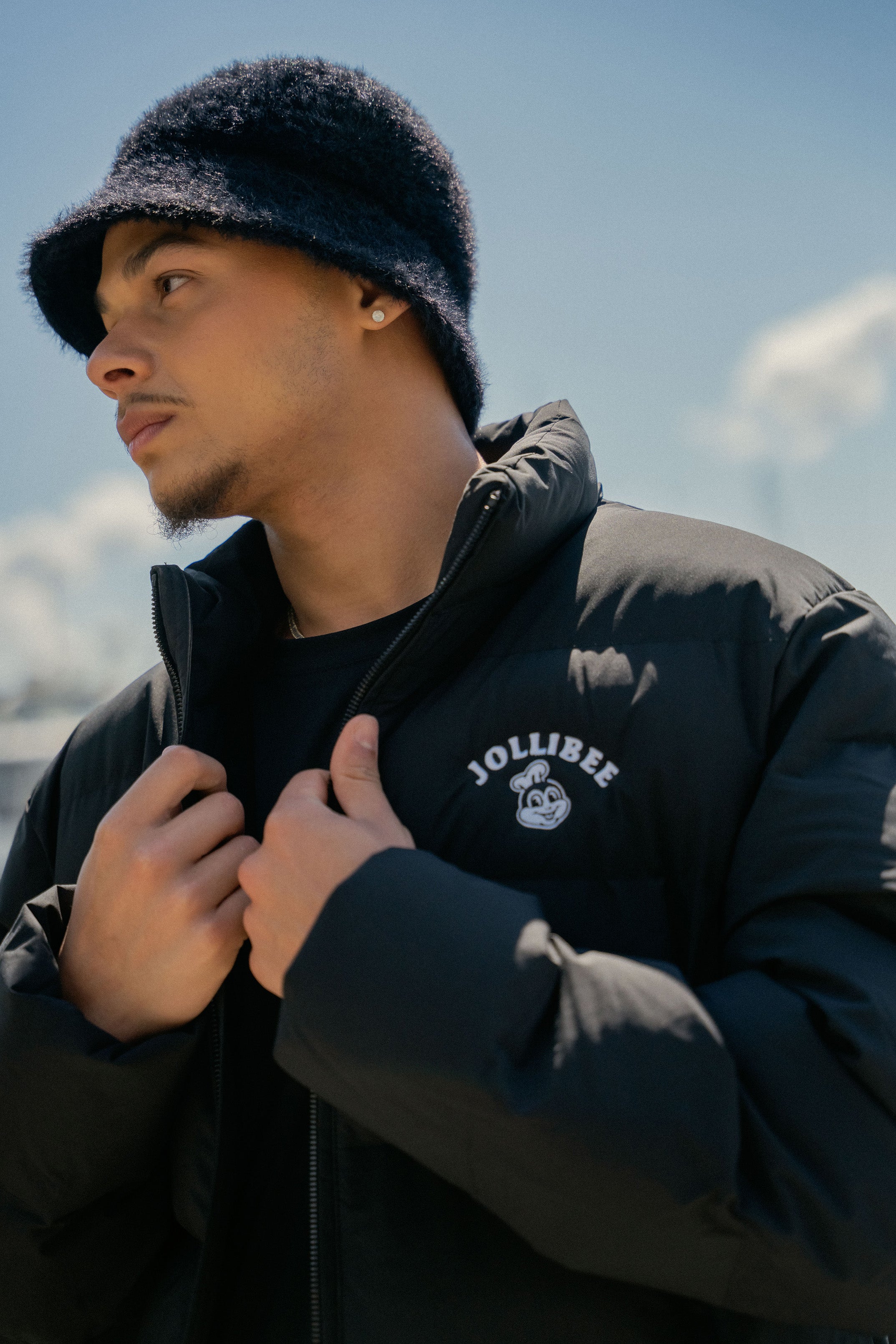Person wearing a black jacket with 'Jollibee' branding against a cloudy sky.