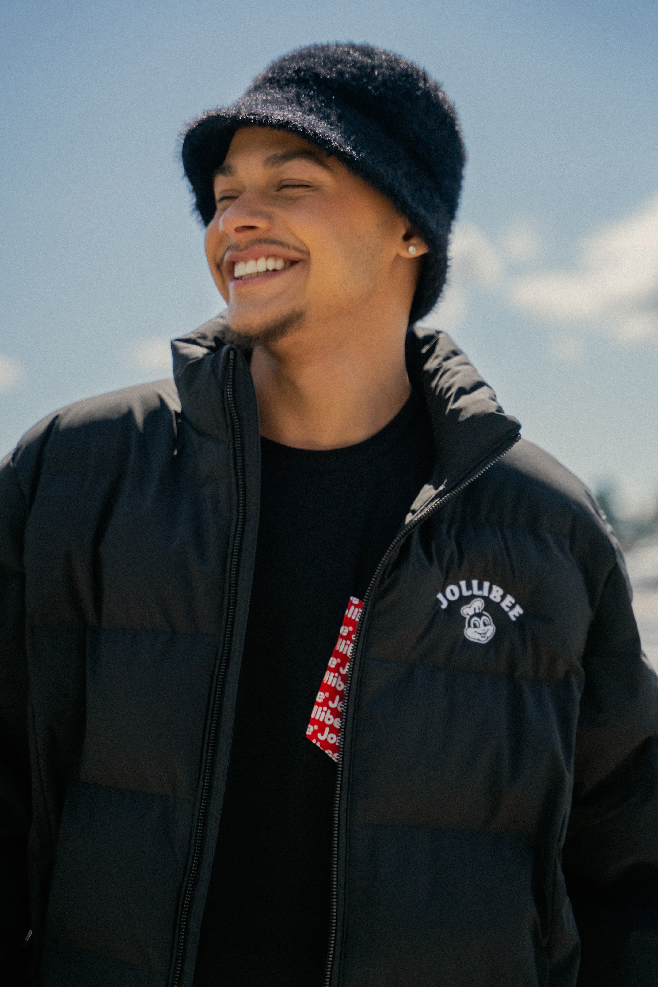 Man wearing a black jacket with a visible Jollibee brand logo, smiling outdoors.