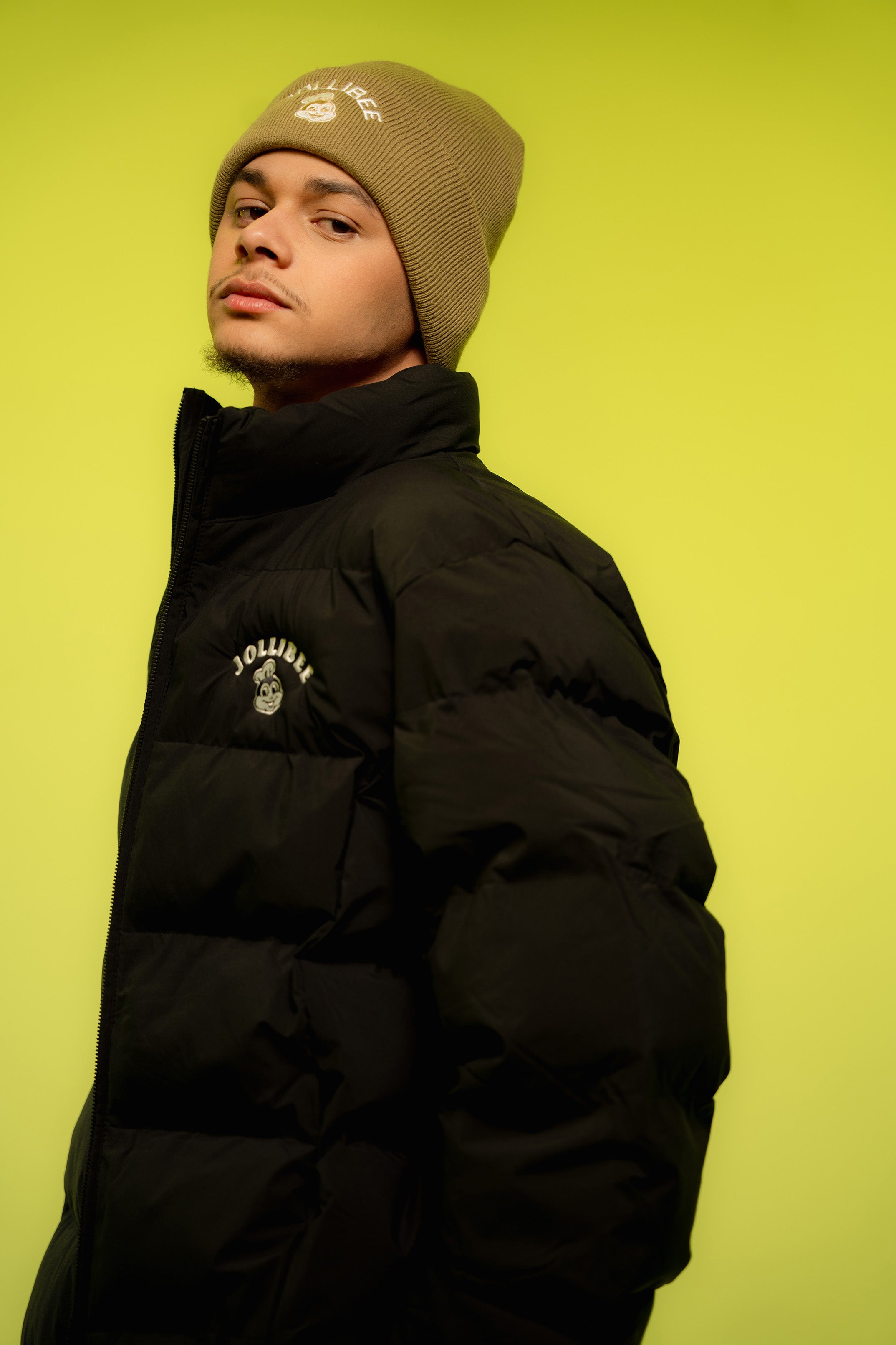 Person wearing a black puffer jacket and beige beanie against a bright yellow background