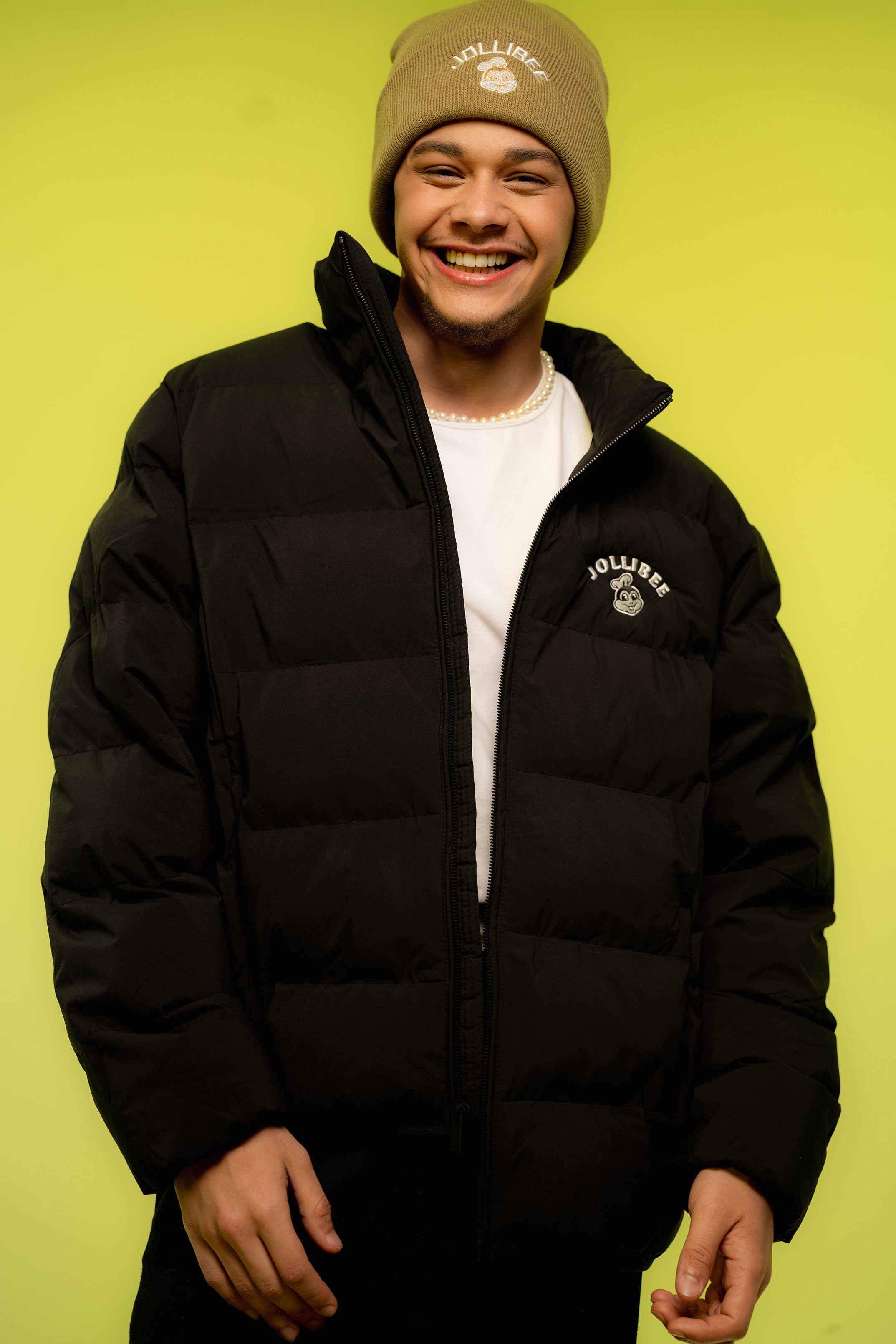 Person wearing a black puffer jacket and beige beanie with a logo on a green background