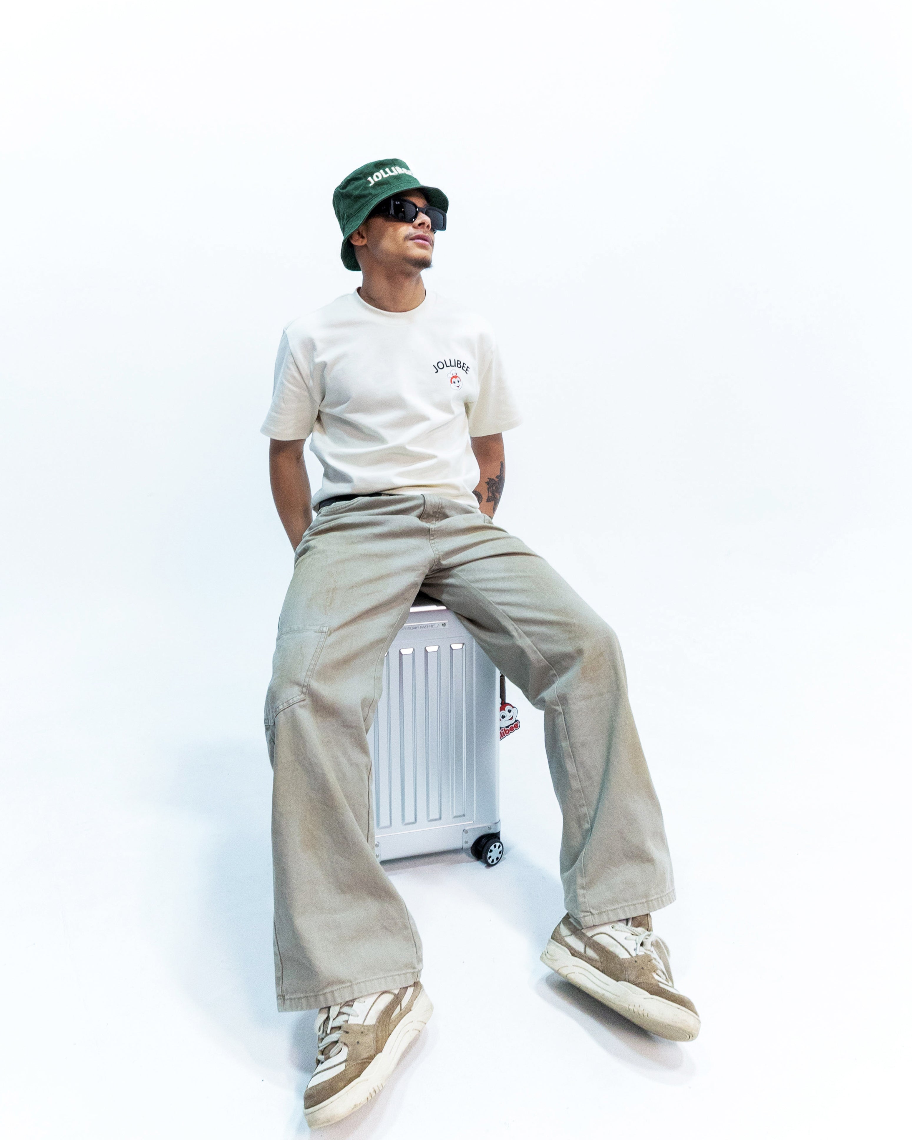 Person wearing a white t-shirt, beige pants, and a green cap sitting on a white box with a white background.