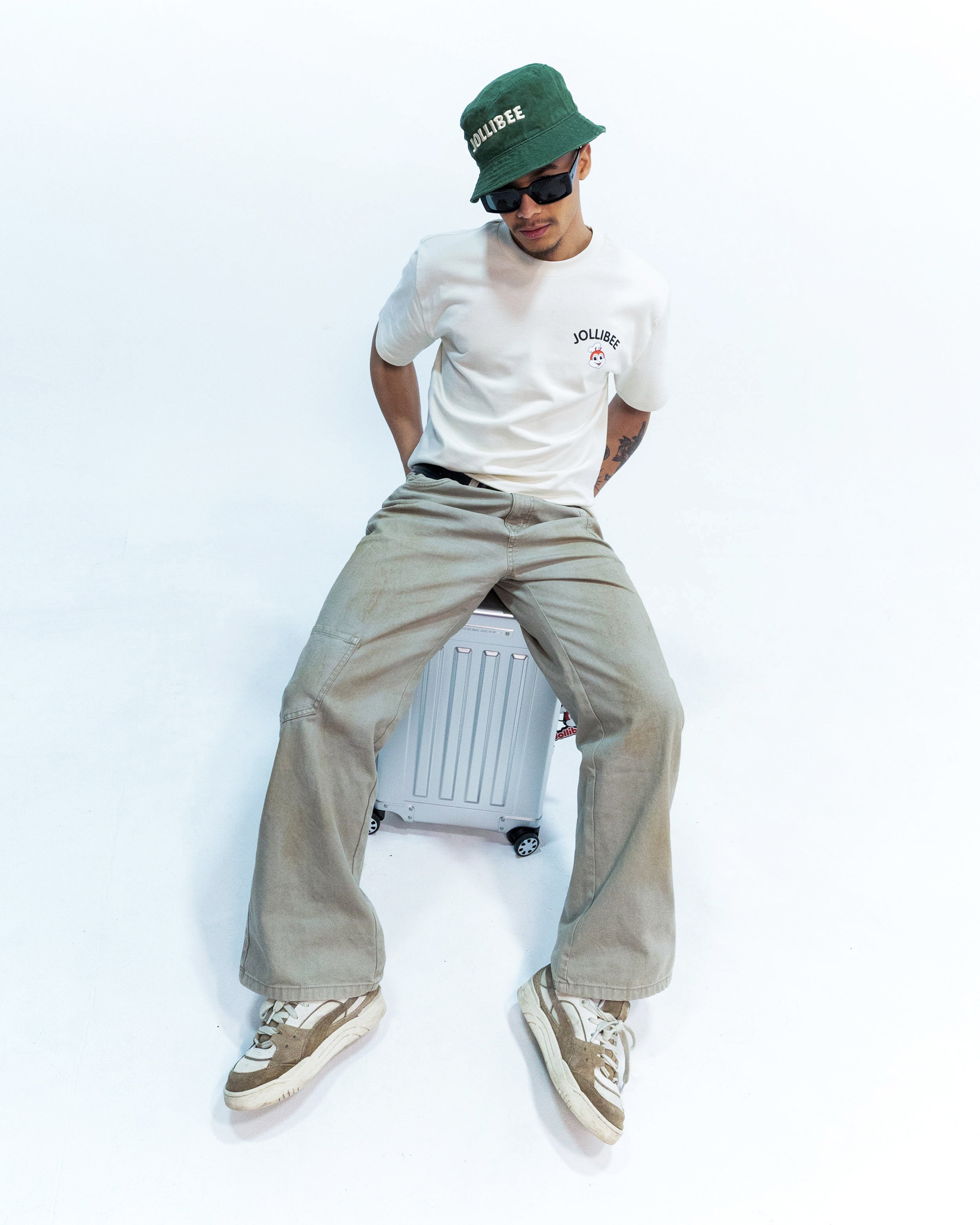 Person sitting on a luggage wearing a Jollibee branded white t-shirt, beige pants, and a green cap with sunglasses.