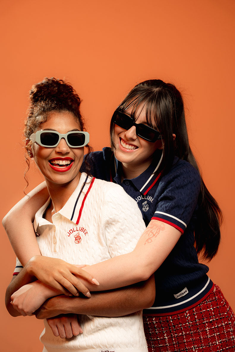 Two people posing together against an orange background, wearing sunglasses and matching Jollibee branded cable knit polos.