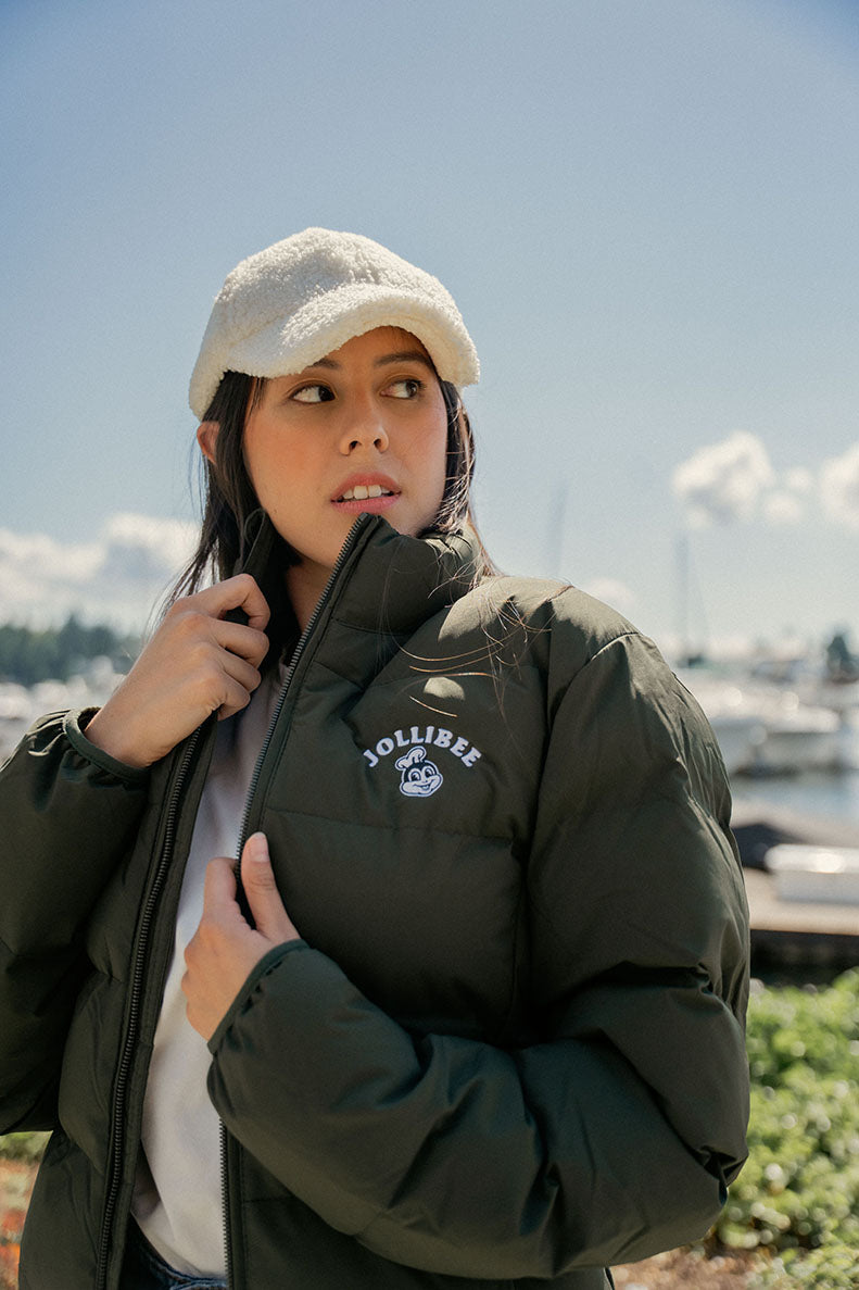 Person wearing a green jacket with a Jollibee logo, white cap, and white shirt outdoors.