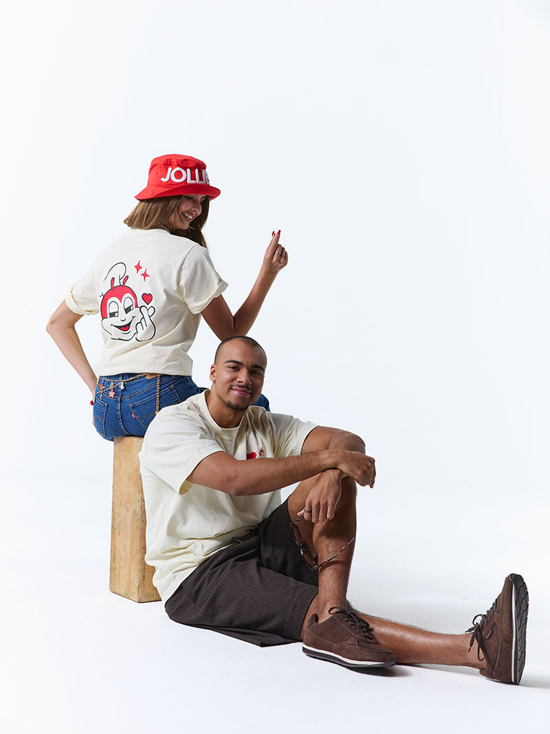 Two people posing against a white background, one sitting and one standing, wearing casual clothing.