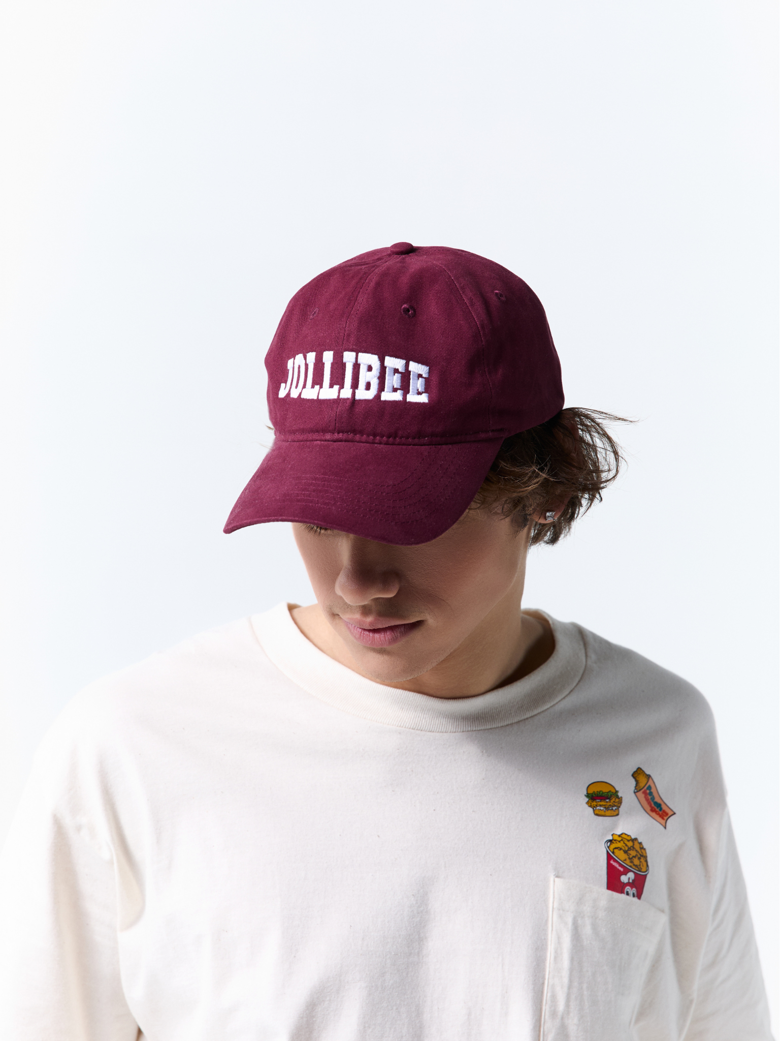 A person wearing a maroon baseball cap with 'JOLLIBEE' embroidered in white on the front, paired with a white long-sleeve shirt against a plain white background.