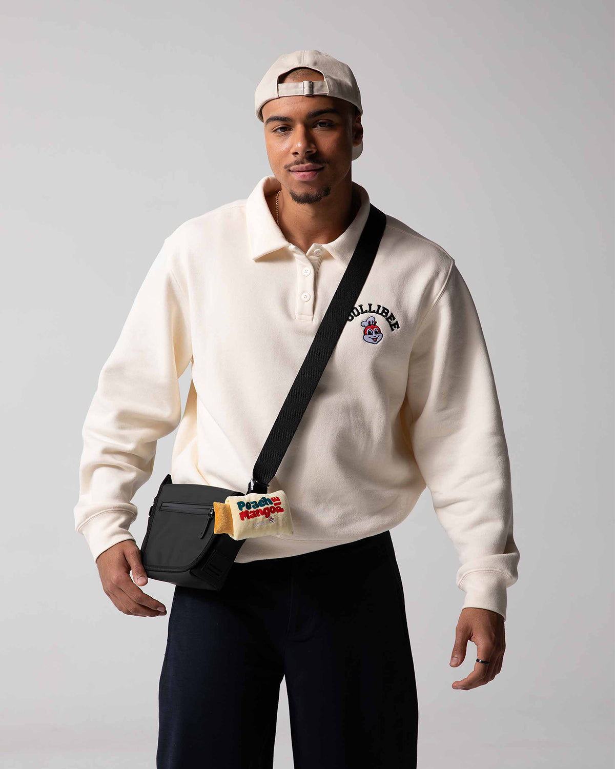 Man wearing a beige sweatshirt with a Jollibee logo, dark pants, and a black crossbody bag on a gray background