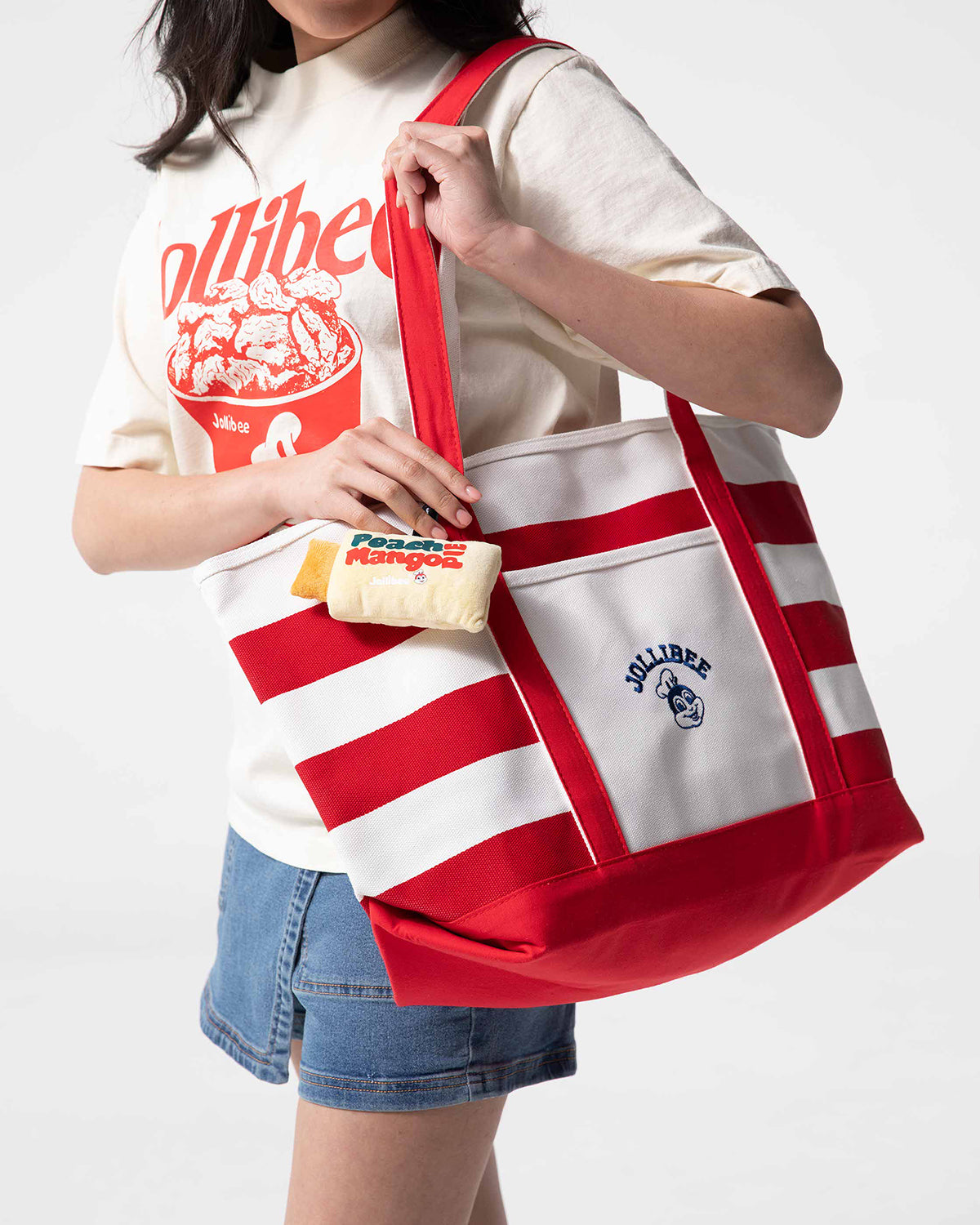 Person wearing a beige t-shirt with red graphics and holding a red and white bag with a Peach Mango Pie plush charm.