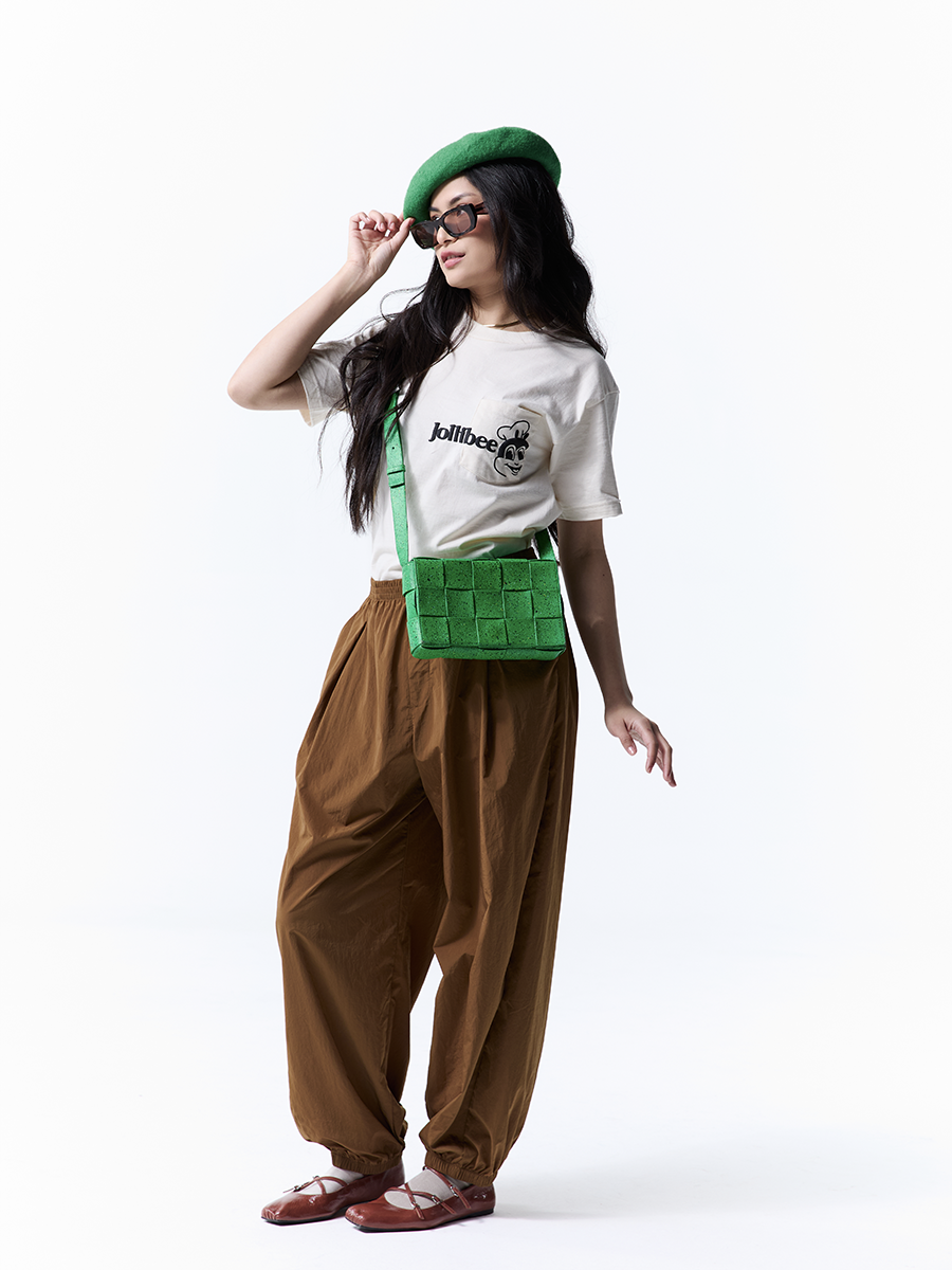 Person wearing a beige t-shirt with Jollibee logo, brown pants, and a green beret and bag on a white background