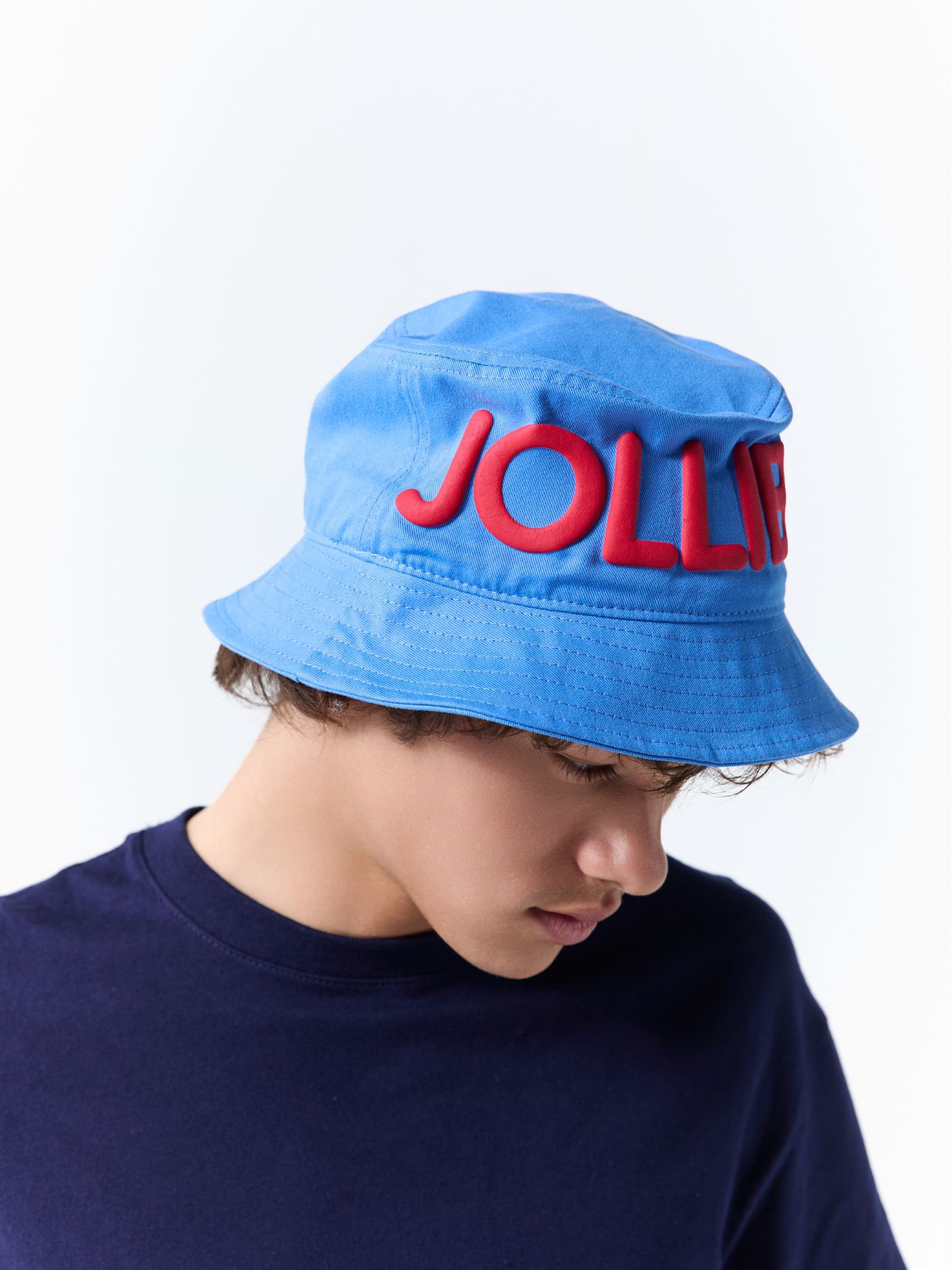 A person wearing a bright blue bucket hat with large red 3D lettering that reads 'JOLLIBEE' across the front, paired with a dark navy crewneck shirt, against a plain white background.