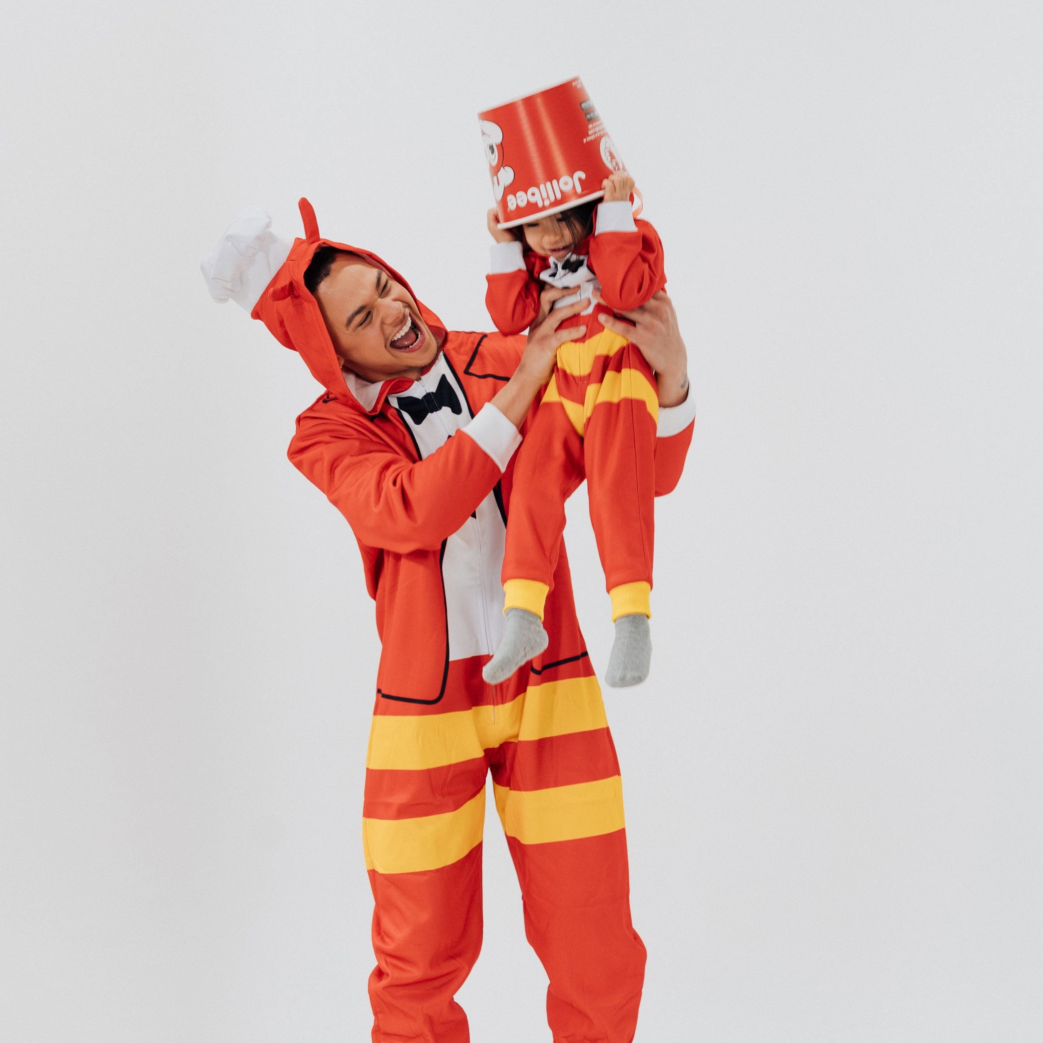 Person wearing a red and yellow Jollibee onesie with a child also in a similar outfit on a white background